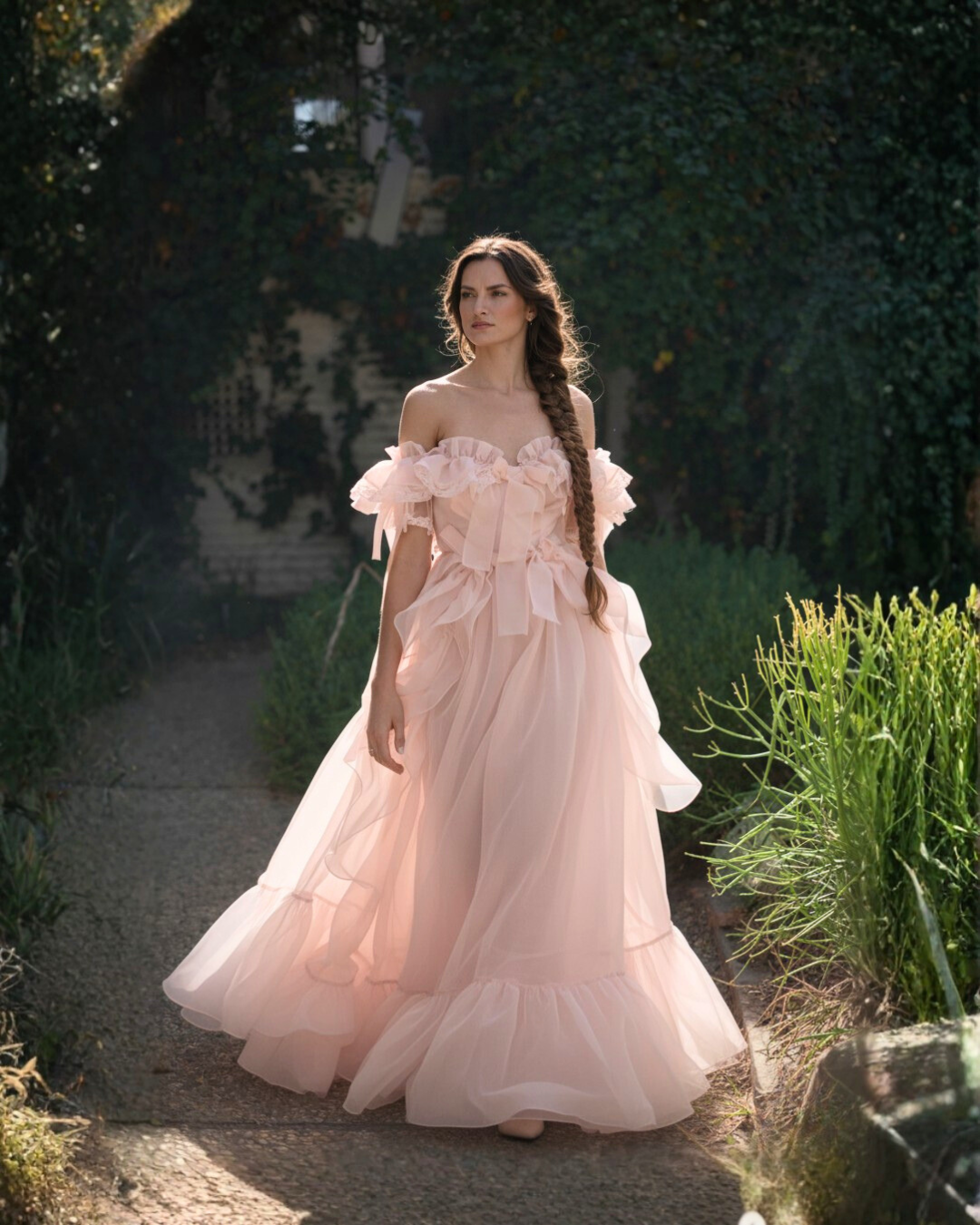 A woman in a flowing, off-shoulder pink dress with ruffle and bow details, standing on a garden path surrounded by greenery.