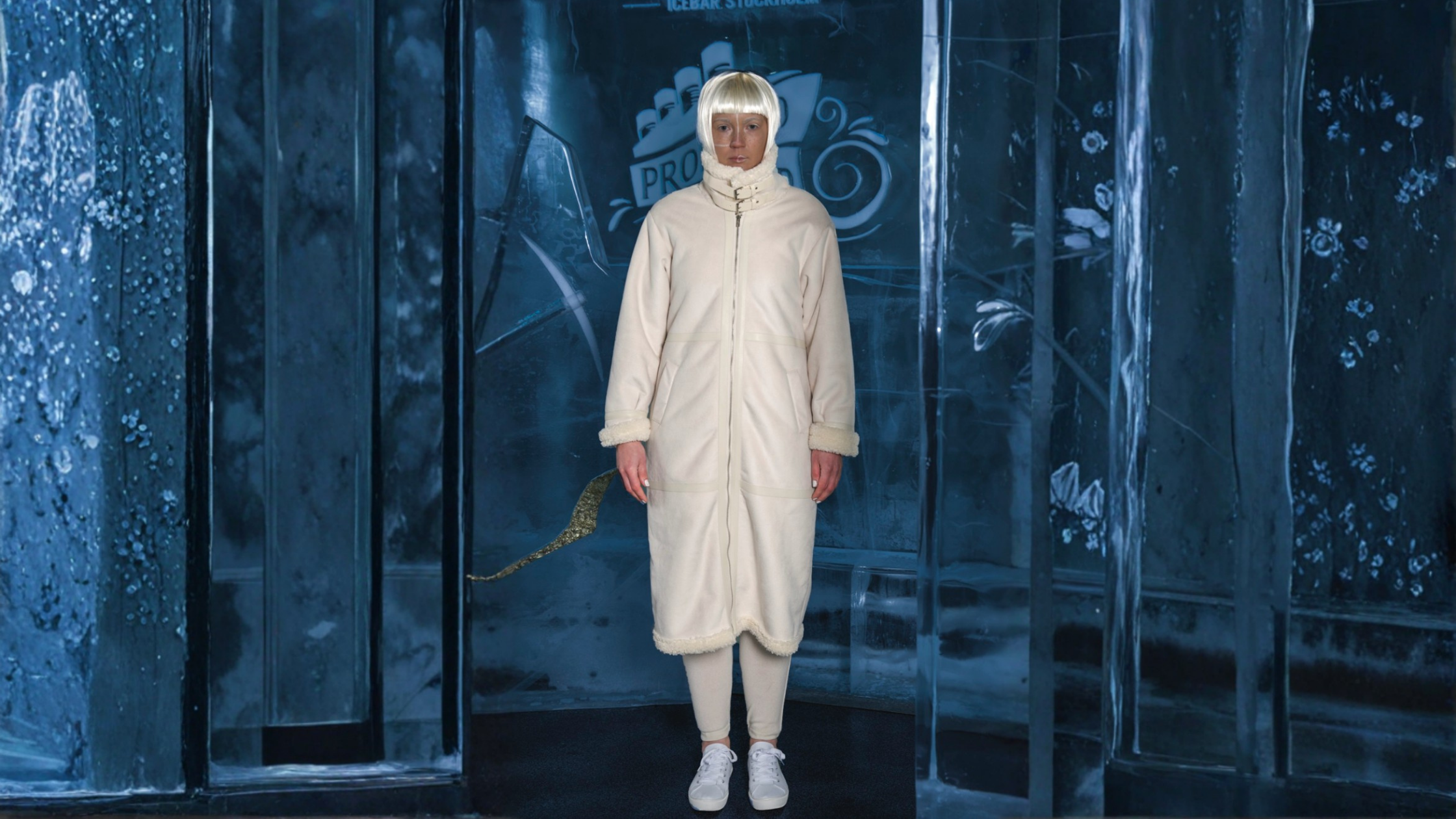 A person stands inside an ice-themed room wearing a long white coat, white sneakers, and white pants. They have a short platinum blonde wig. The background features icy walls with snowflakes and a sign that reads 'PRO'.