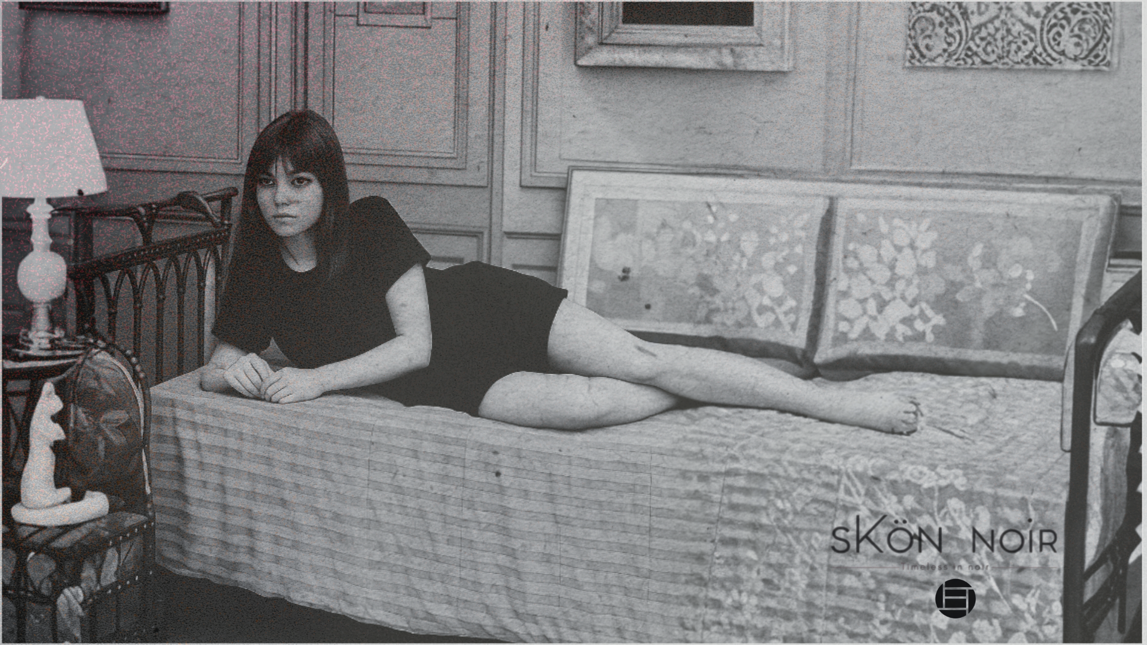 A woman with dark hair in a black outfit lying on a bed in a vintage room with paneled walls and framed artwork, next to a lit table lamp and a small decorative chair.