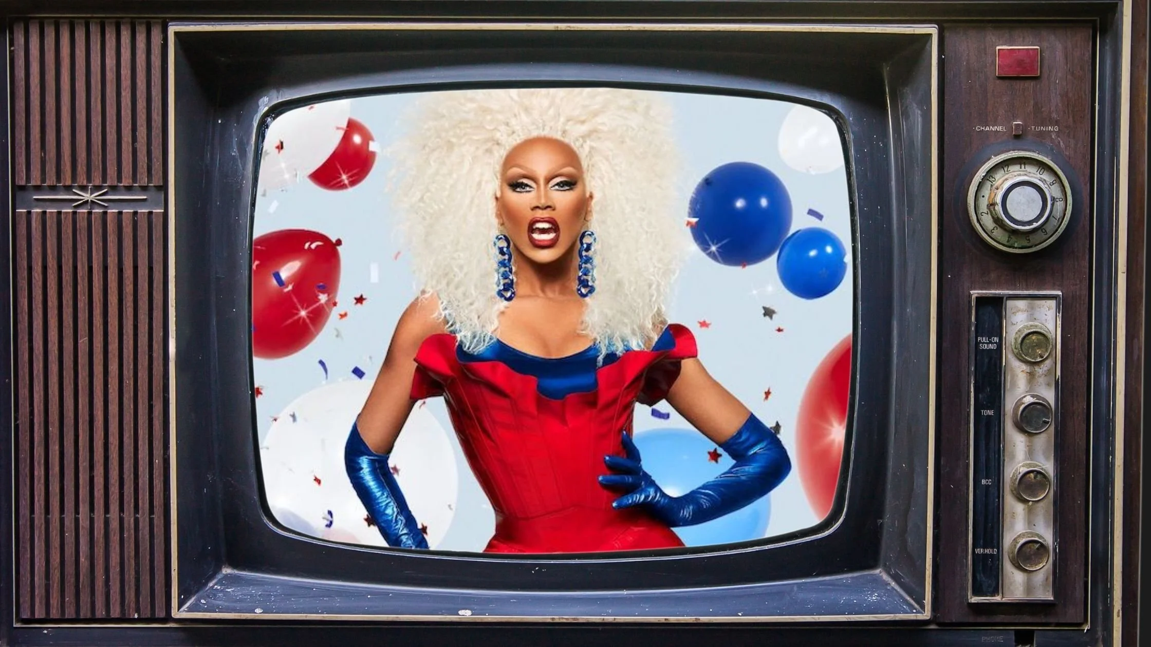 A person dressed in a vibrant red and blue fantasy costume with large blue earrings, standing inside an old-fashioned TV set with balloons and confetti in the background.