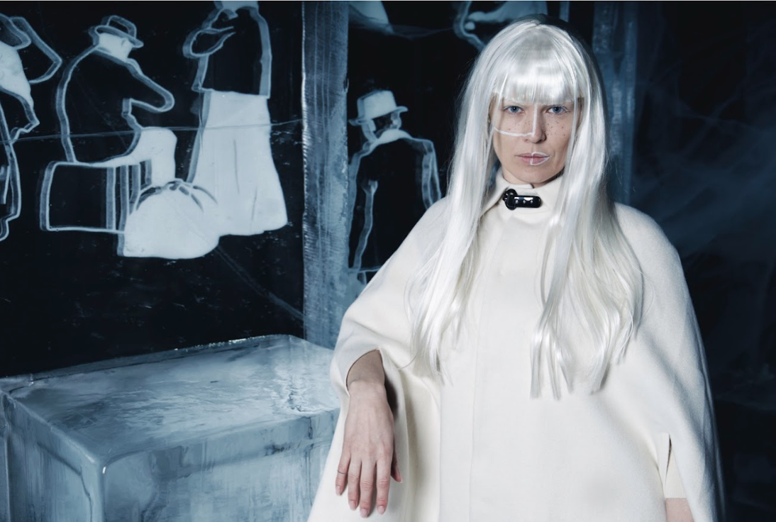 Person with long white hair, wearing a white cape and glasses, standing in front of a black wall with white outline drawings of people wearing hats.