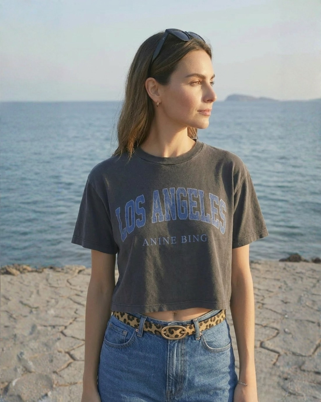 A young woman with light skin, shoulder-length brown hair, and sunglasses on her head stands on a rocky beach near the ocean, looking away from the camera.