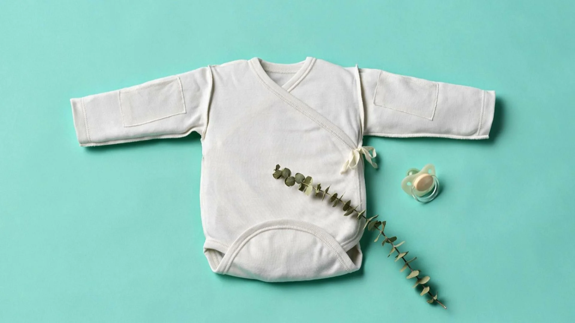 A white baby onesie with long sleeves laid flat on a light blue background, accompanied by a pacifier and a sprig of eucalyptus.
