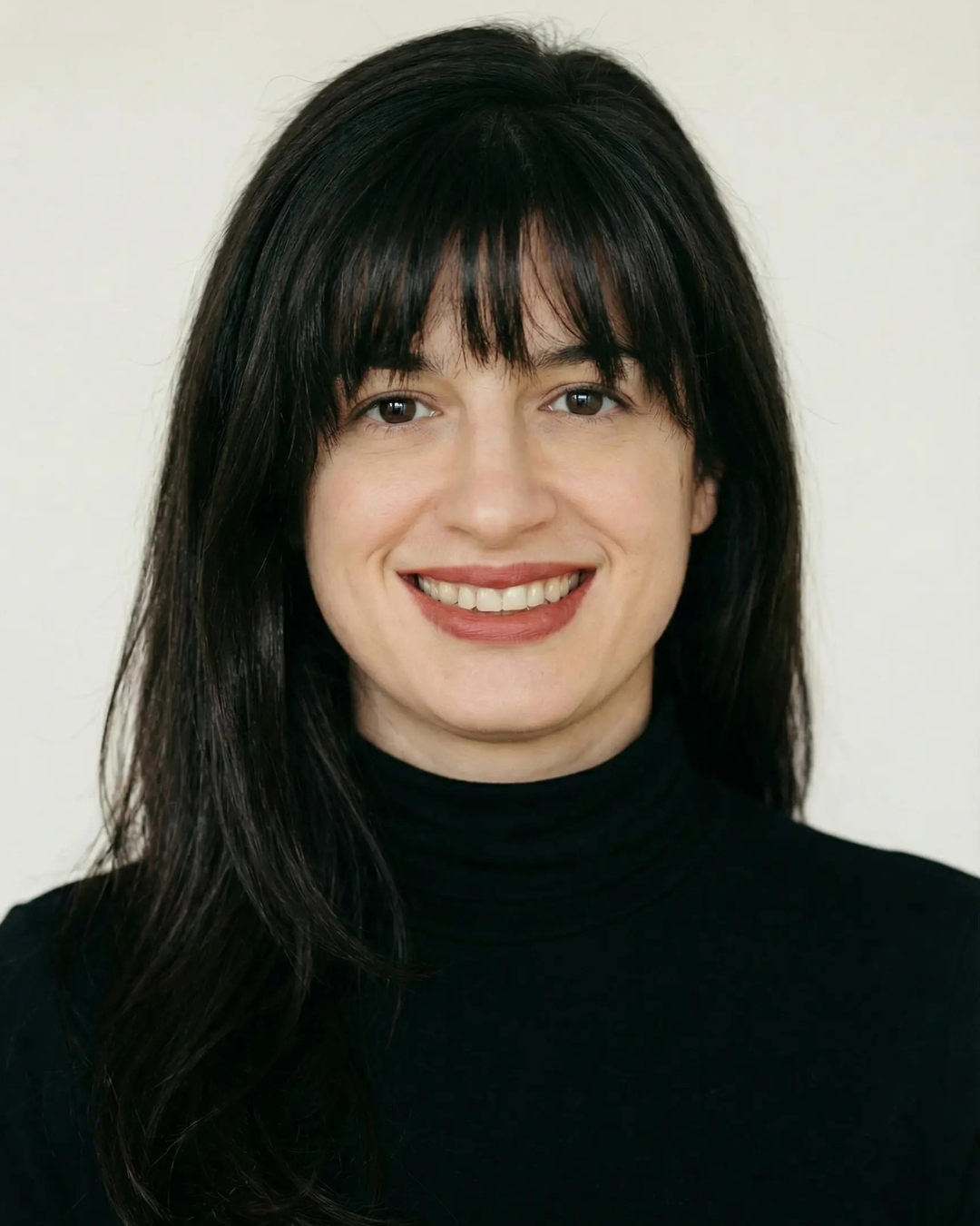 A woman with long dark hair and bangs, wearing a black turtleneck, smiling in front of a plain light background.