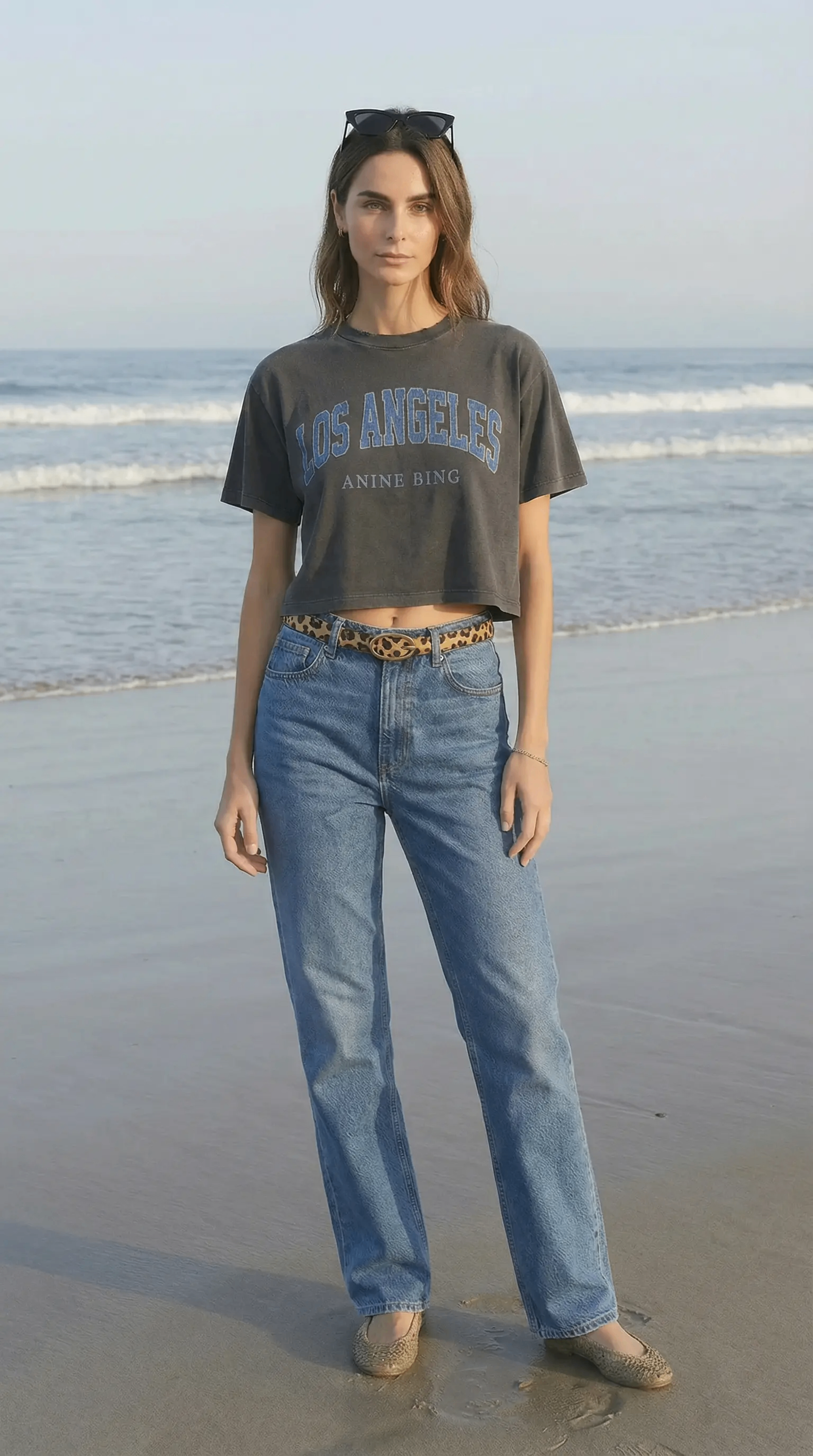 A young woman standing on the beach near the water, wearing a cropped T-shirt with 'Los Angeles' text, high-waisted jeans, leopard print belt, and sunglasses on her head.