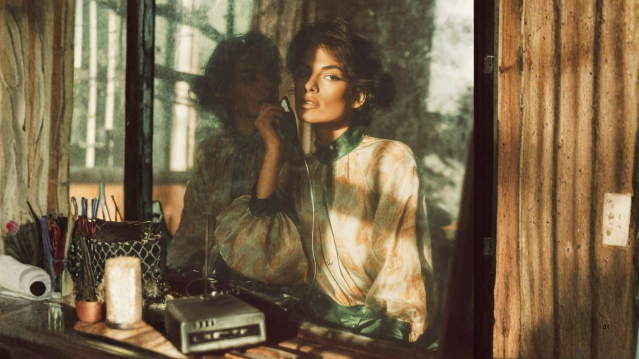 A woman with short, dark hair and makeup, wearing a silky, patterned blouse, is talking on a vintage telephone. She is looking into a window, and her reflection is visible on the glass. The scene appears to be inside a rustic wooden room with various items on the table in the foreground.