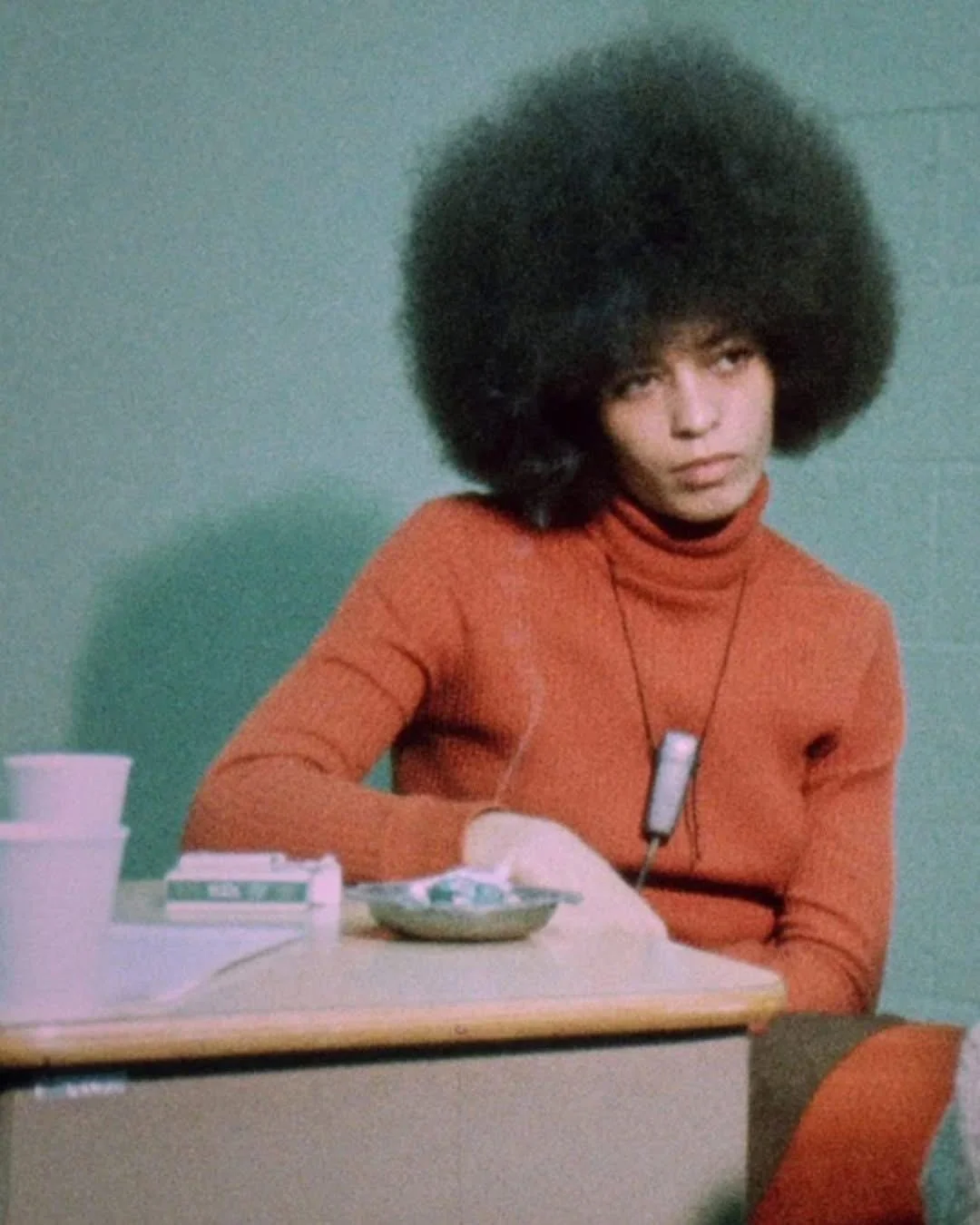 A young woman with a large afro hairstyle sitting at a desk with a serious expression, wearing an orange turtleneck sweater and a necklace with a rectangular pendant.
