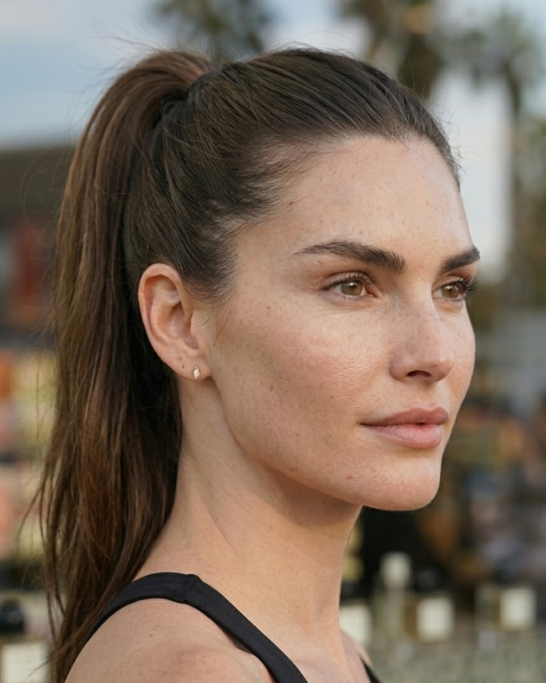 Close-up of a woman with long brown hair tied in a high ponytail, wearing small stud earrings, looking slightly to the right, outdoors with blurred background.