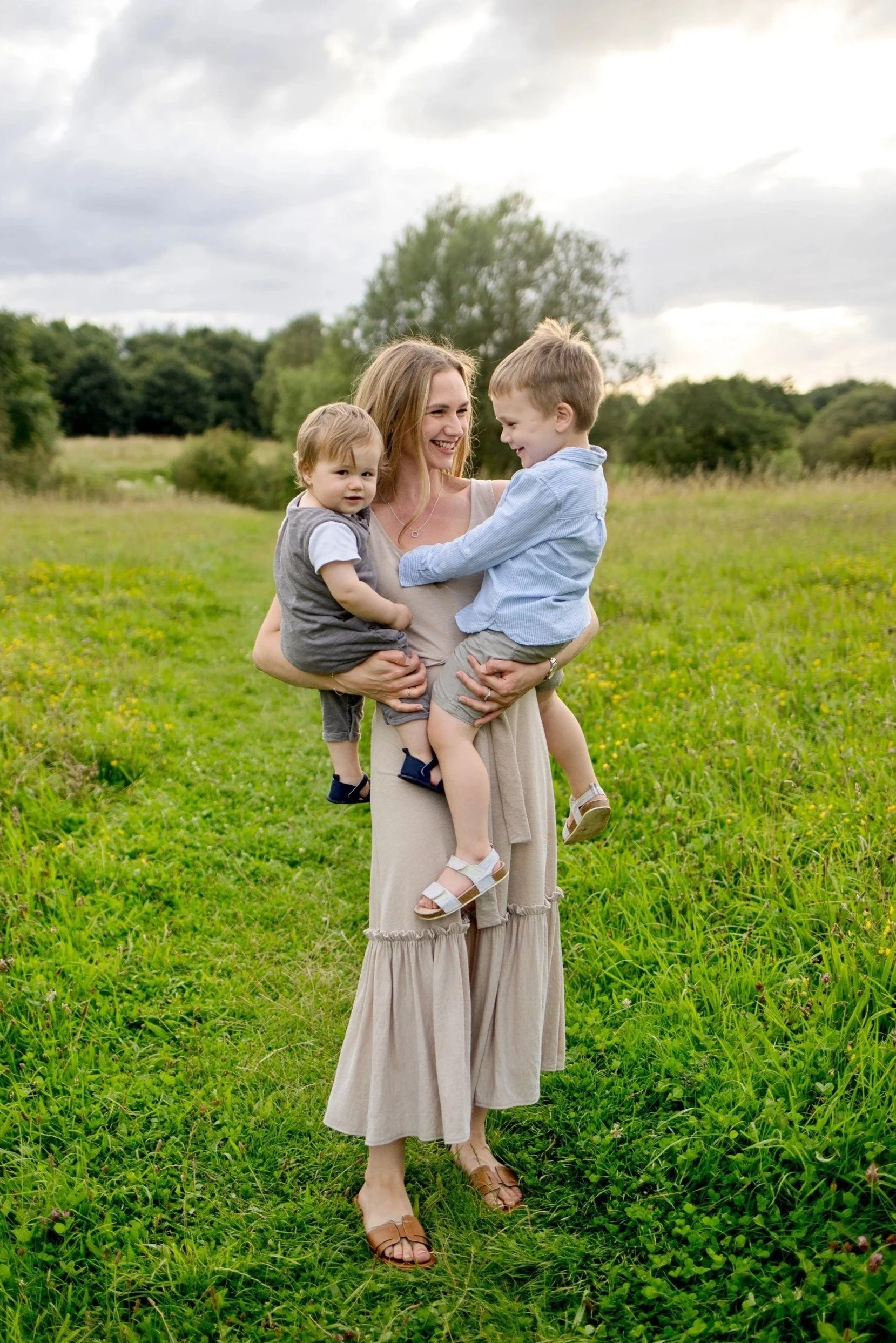 A working mum holding two young children in a grassy field with trees.