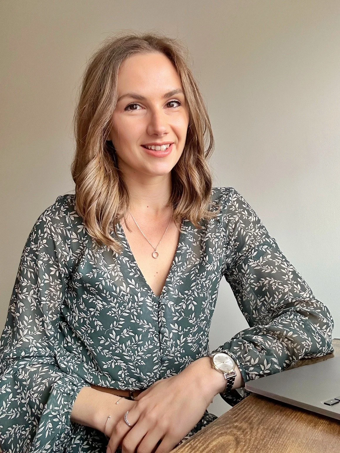 A working mum with wavy brown hair, wearing a floral patterned blouse, sitting at a desk with a laptop. She is smiling confidently as she has found balance from burnout in her approach to parenting, her career and homelife.
