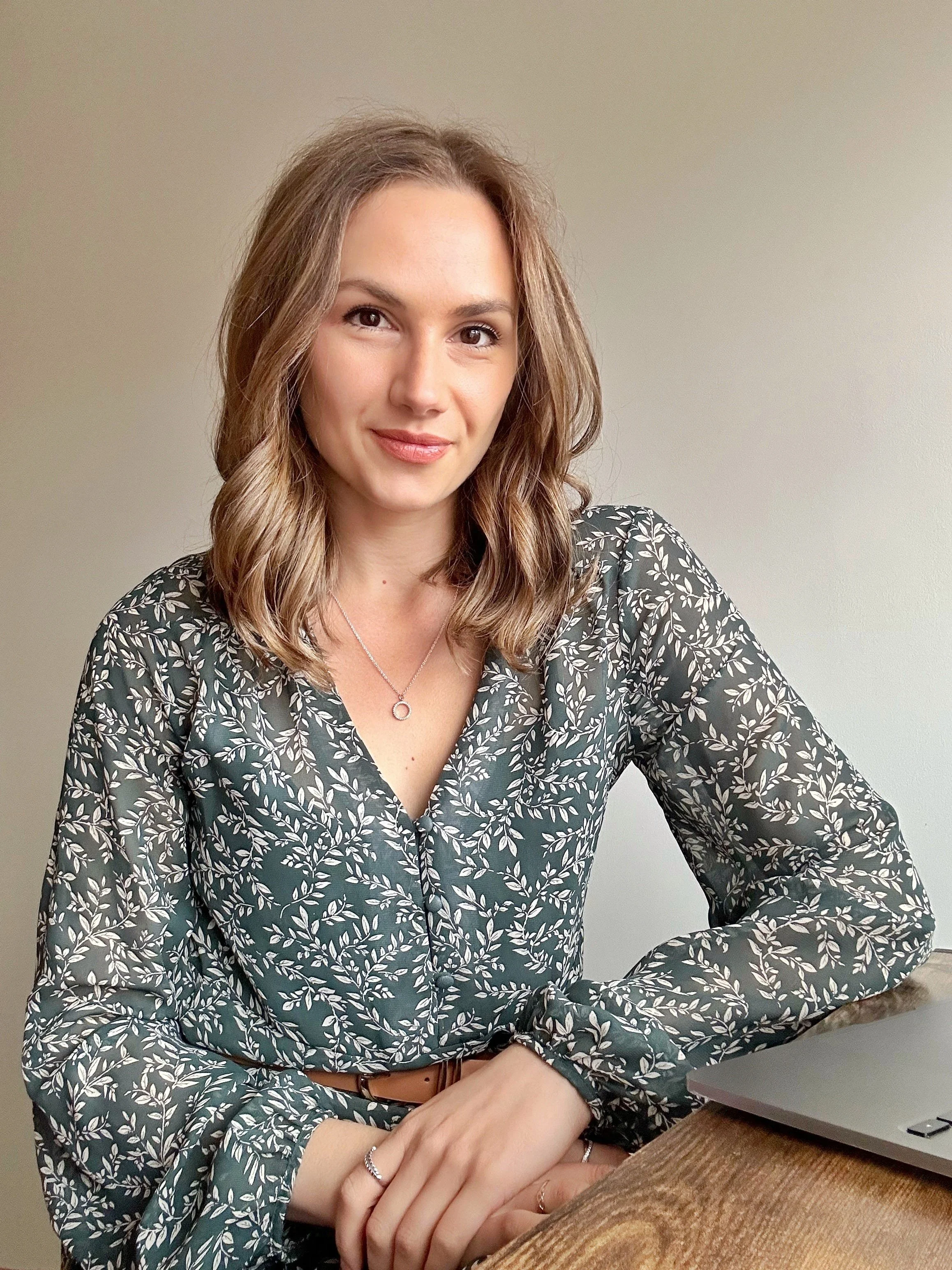 A working mum with wavy brown hair, wearing a floral patterned blouse, sitting at a desk with a laptop. She is smiling confidently as she has found balance from burnout in her approach to parenting, her career and homelife.