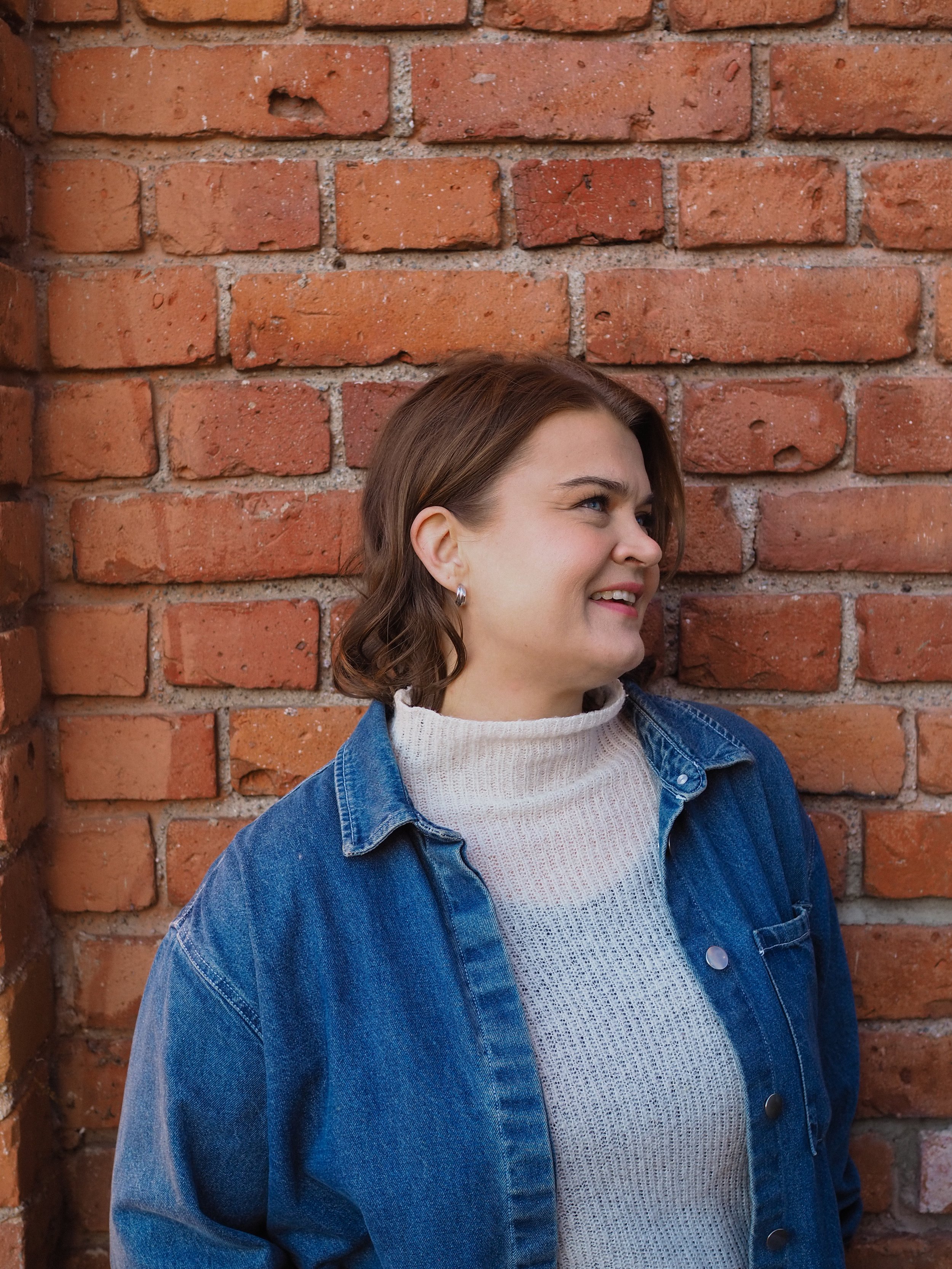 Young woman with short brown hair, wearing a gray turtleneck sweater and a blue denim jacket, smiling and leaning against a brick wall.