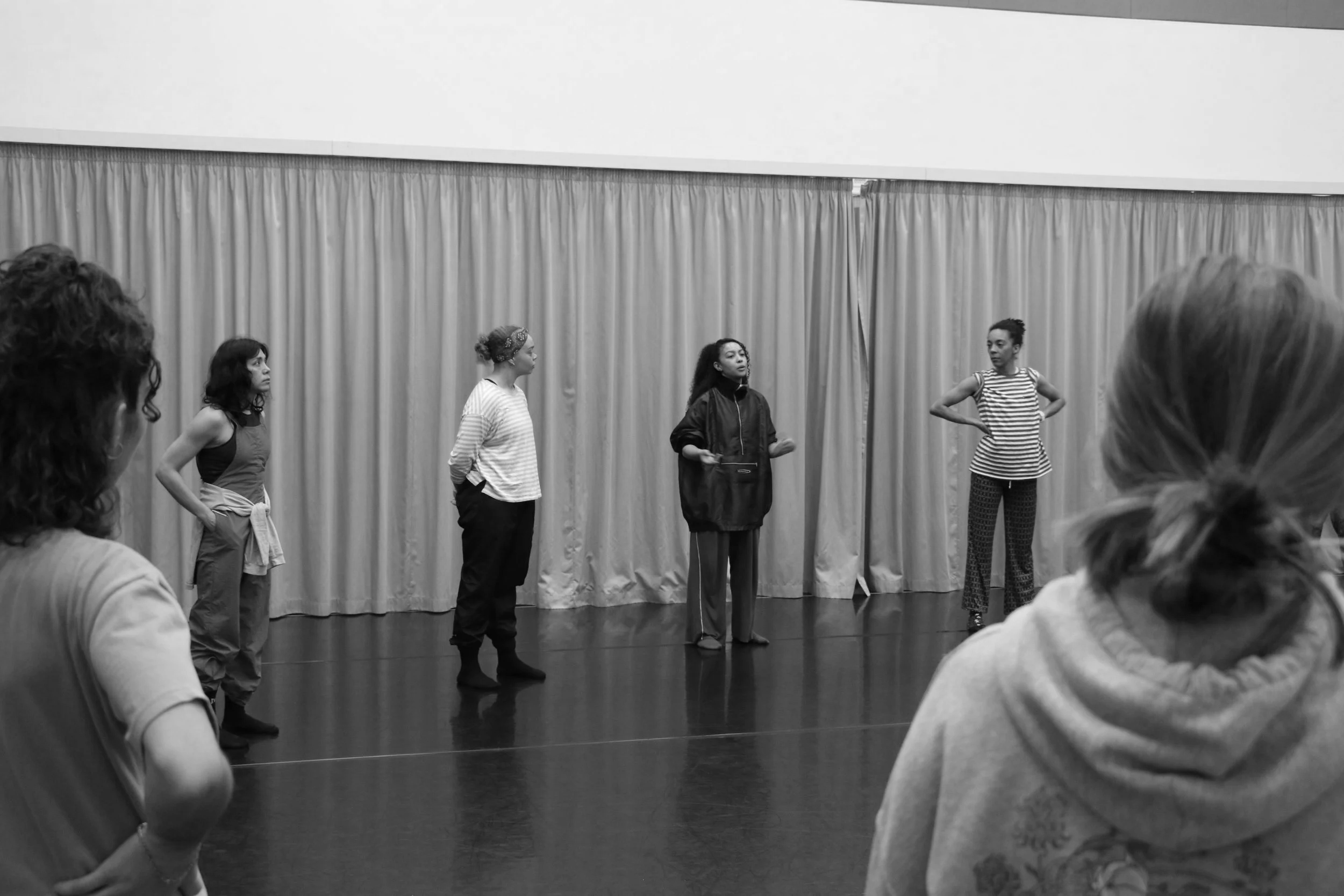 A group of actors or dancers rehearsing on stage in front of a curtain, with several people observing in the foreground.
