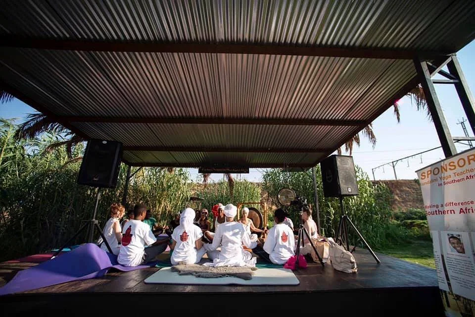 Group of people sitting in a circle on a stage outdoors, with grass and trees in the background, participating in a spiritual or meditation gathering.