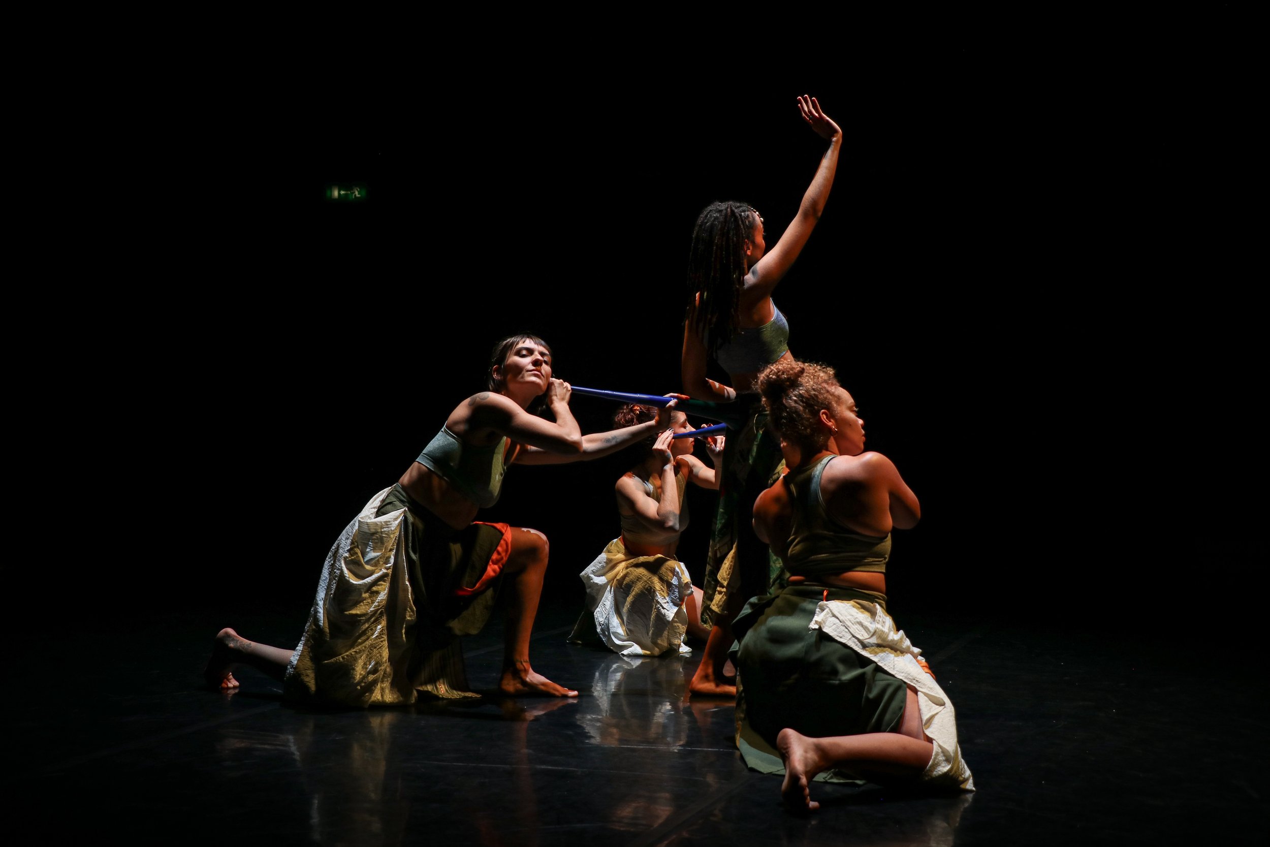 Five dancers perform on a dark stage, with one standing and raising an arm, and the others kneeling or crouching, wearing tribal or cultural costumes.