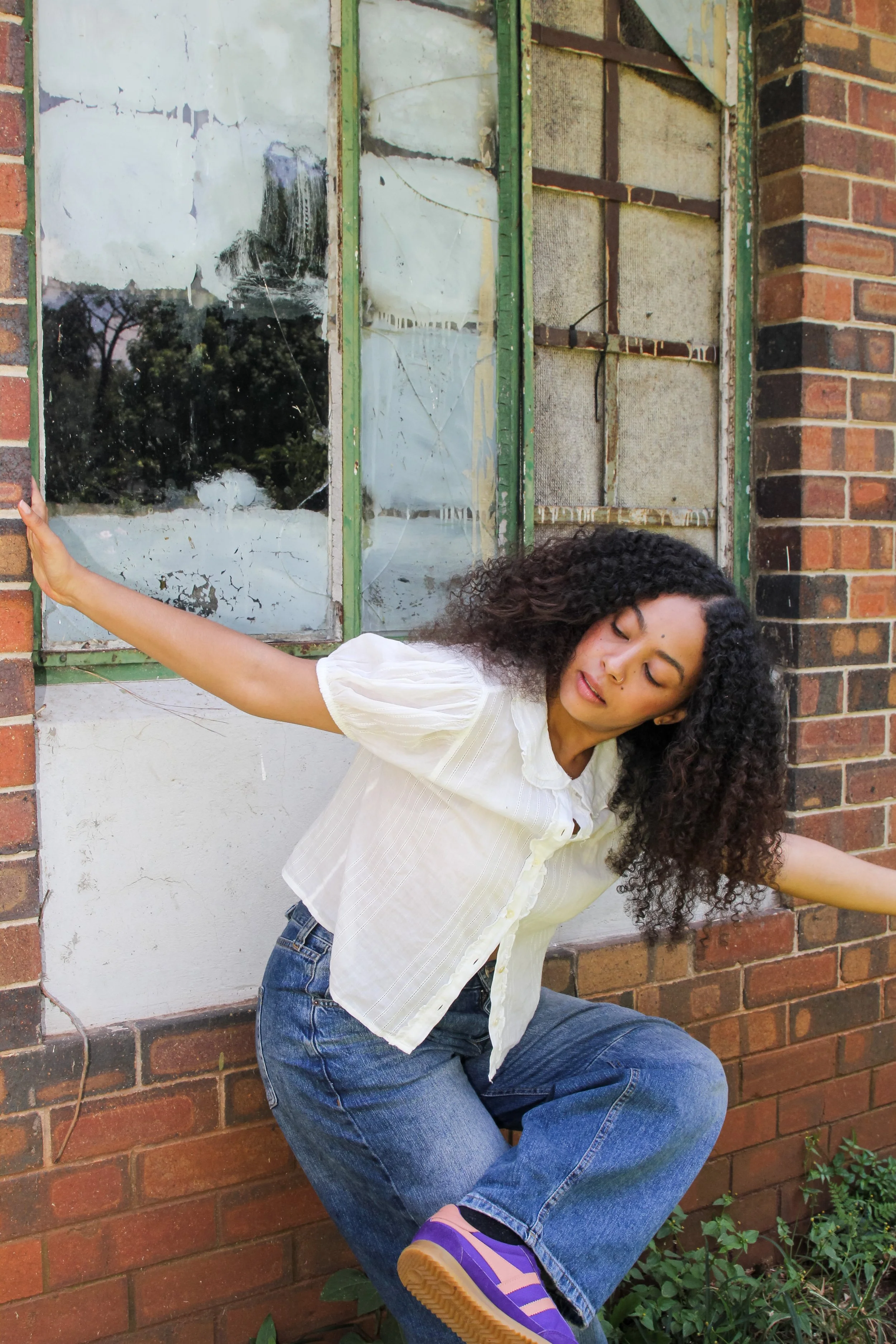 Young woman with curly hair wearing a white blouse and jeans, balancing on one leg with arms outstretched, in front of a dilapidated brick building with broken windows.