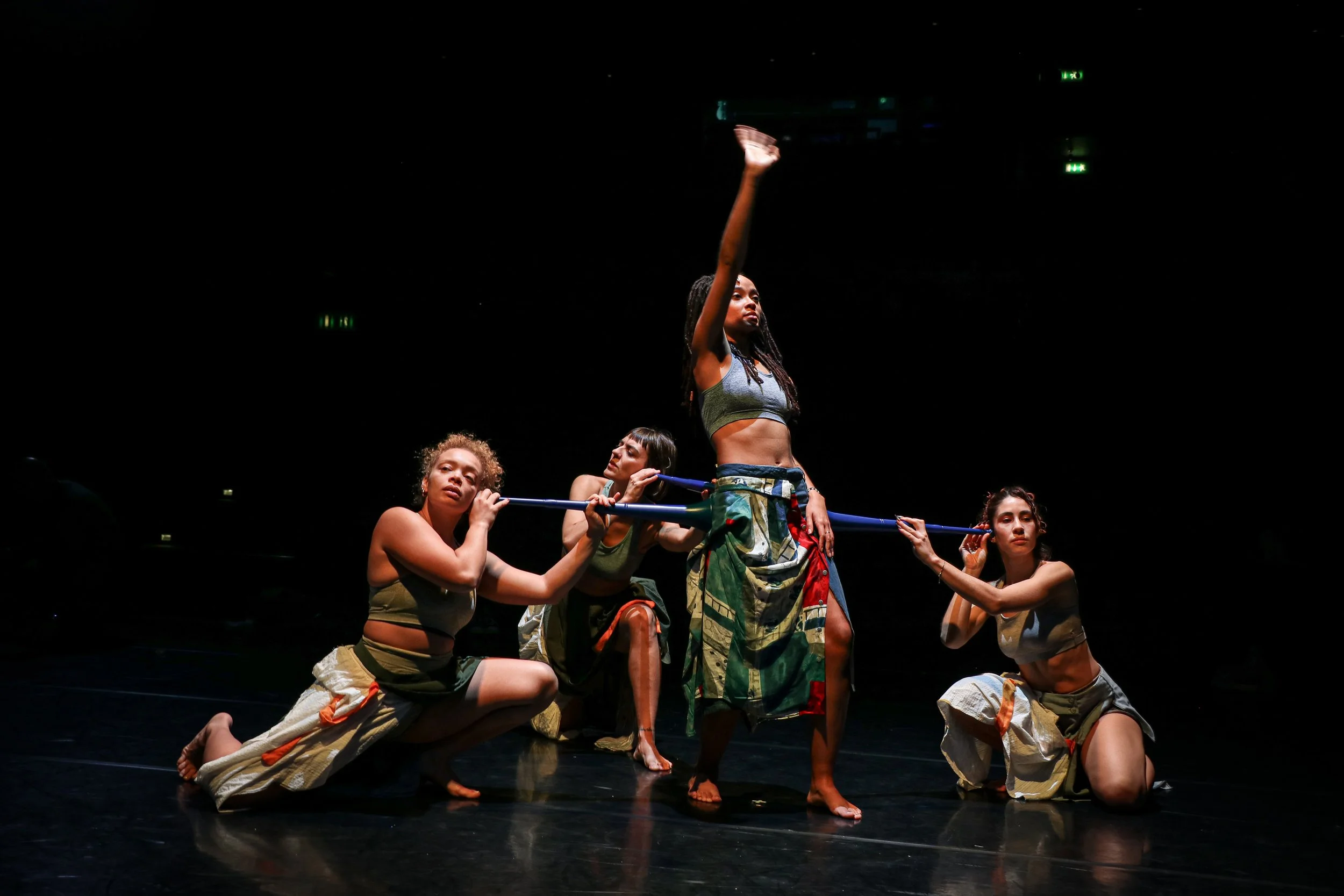 Four women performing a dance or theatrical piece on a dark stage, with one woman standing with her arm raised and the others kneeling or crouched while holding a long stick.