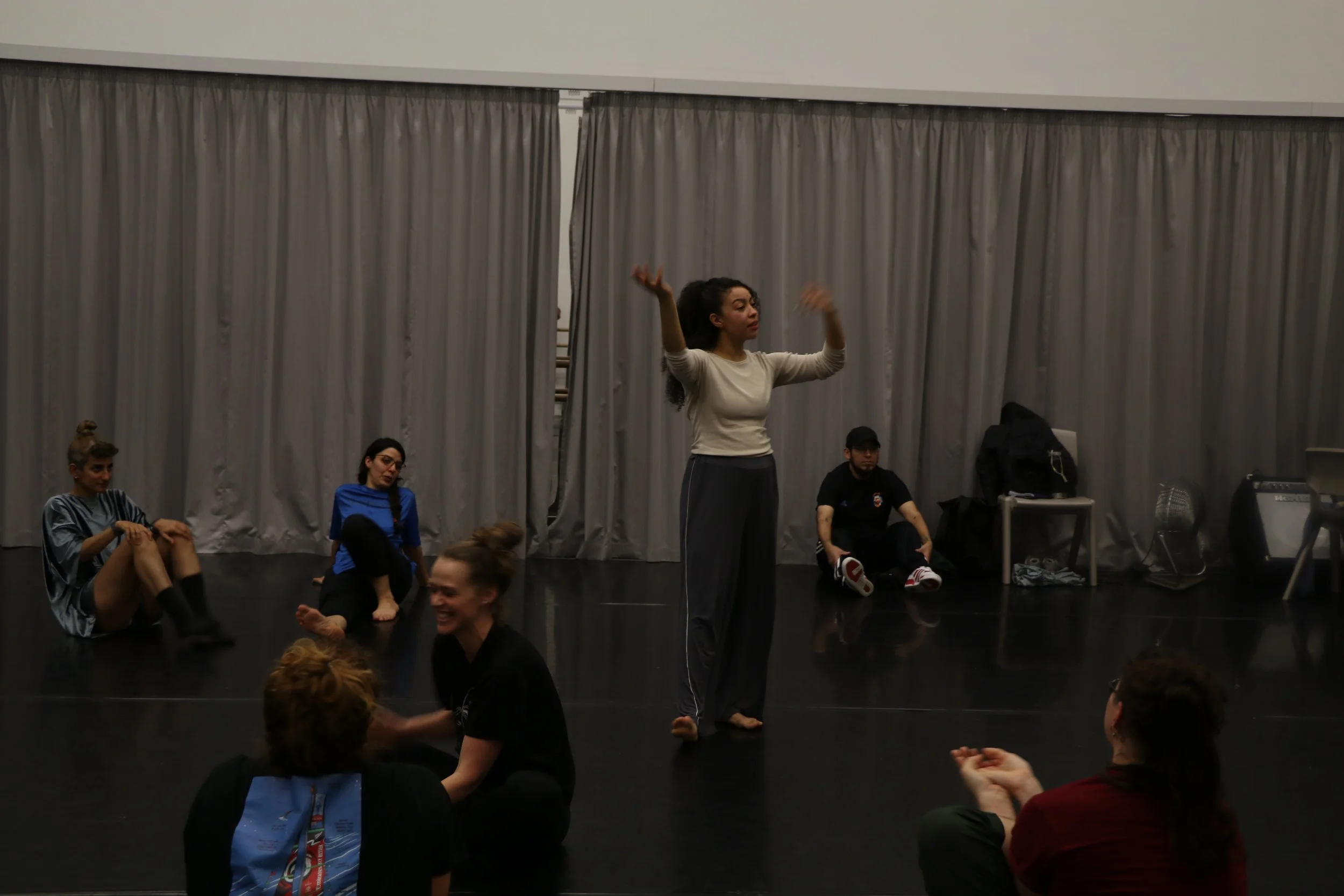 A group of people sitting on the floor in a dance studio, with a woman standing and talking in the center, gesturing with her arms. Others are sitting or lying on the floor, some laughing or smiling. Grey curtains hang in the background.
