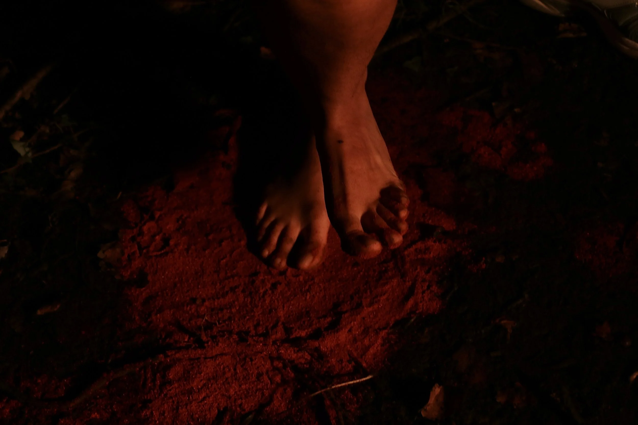 Close-up of bare feet on reddish soil at night, illuminated by a red light.