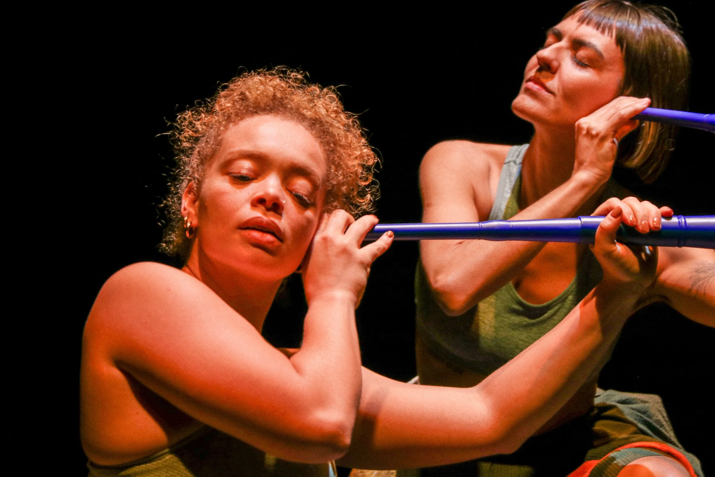 Two women holding blue poles, the woman in the foreground has curly blonde hair and is resting her head on the pole, with eyes closed and a relaxed expression. The woman in the background has straight brown hair, closed eyes, and is leaning on the pole.