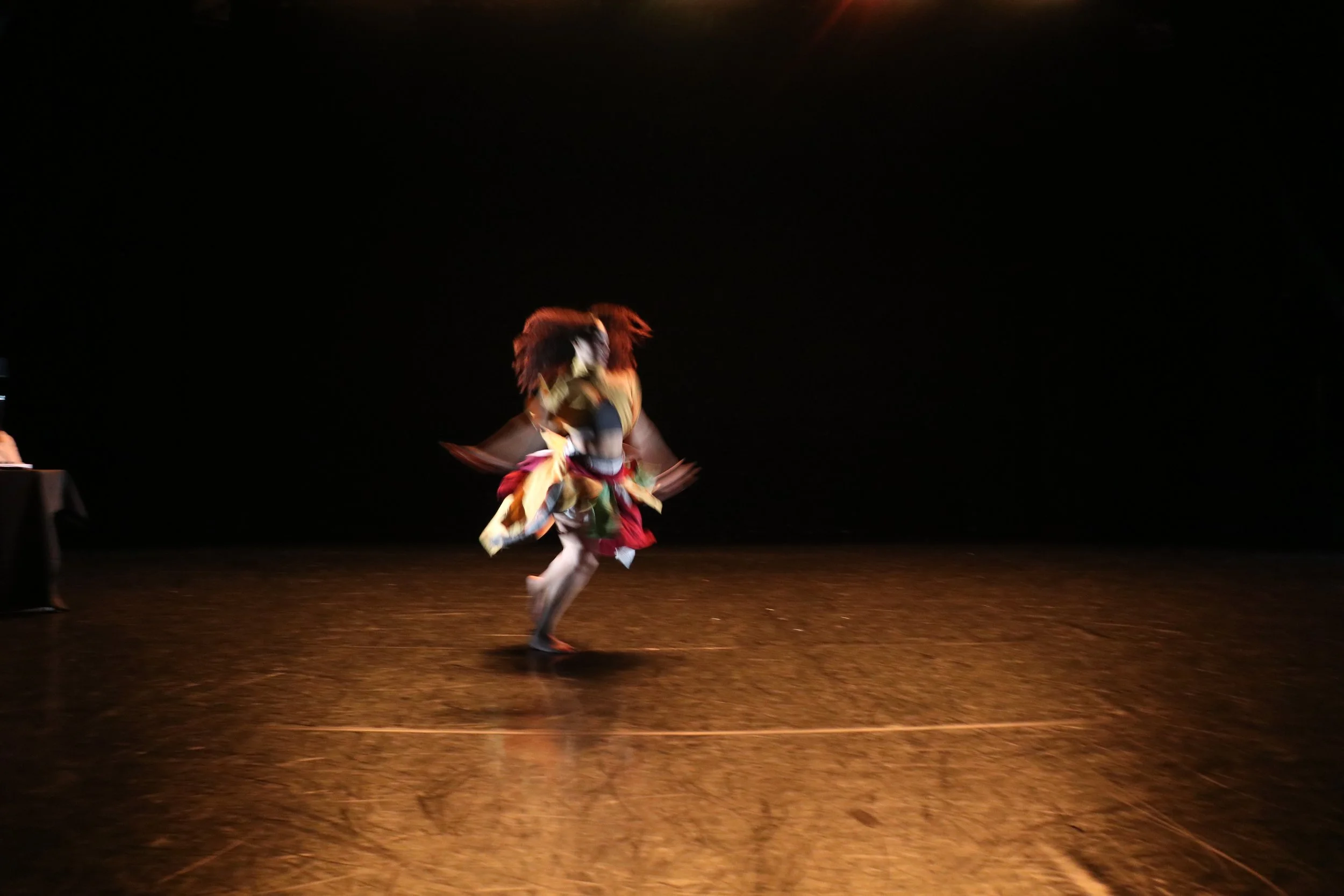 Person in colorful costume performing dance on dark stage with minimal lighting.