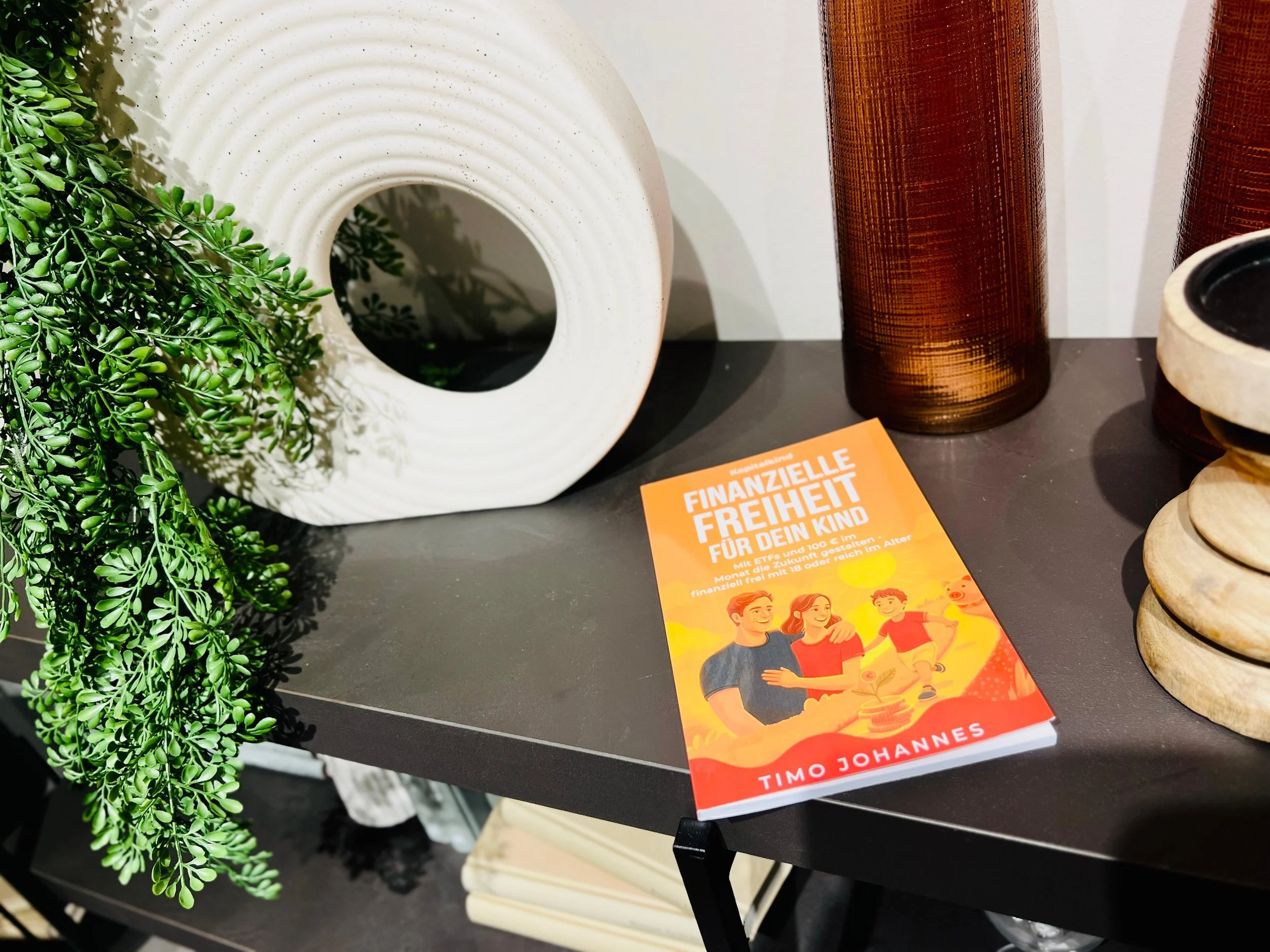 Ein Tisch mit einem orangefarbenen Buch, einer weißen Keramikvase, drei braunen Glasvasen und einem Stack aus runden, beige Keramikschalen. Links eine grüne Pflanze. Das Buch trägt den Titel "Finanzielle Freiheit für dein Kind" von Timo Johannes.