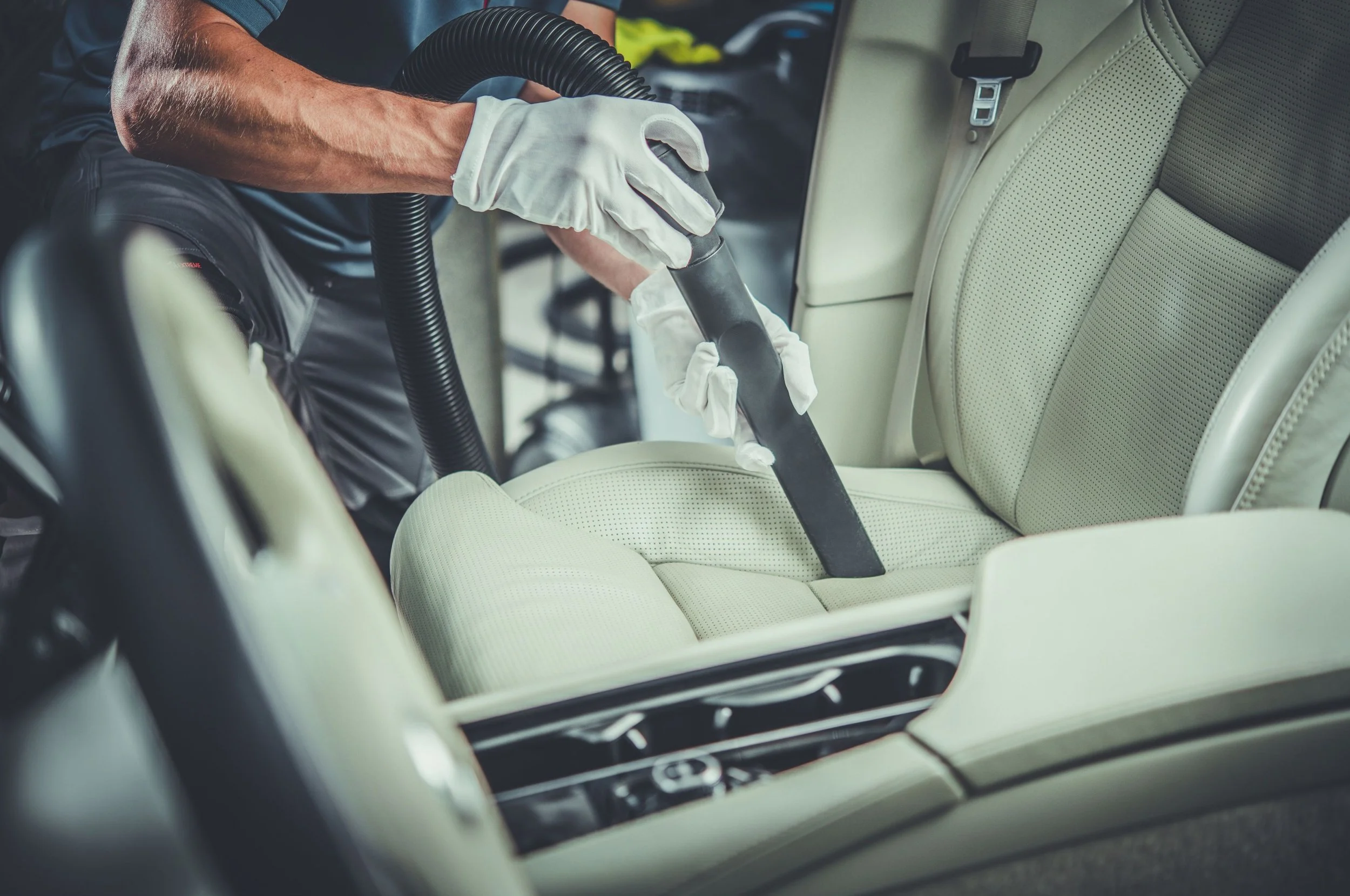 Person vacuuming a beige car seat with a handheld vacuum cleaner, wearing white gloves.