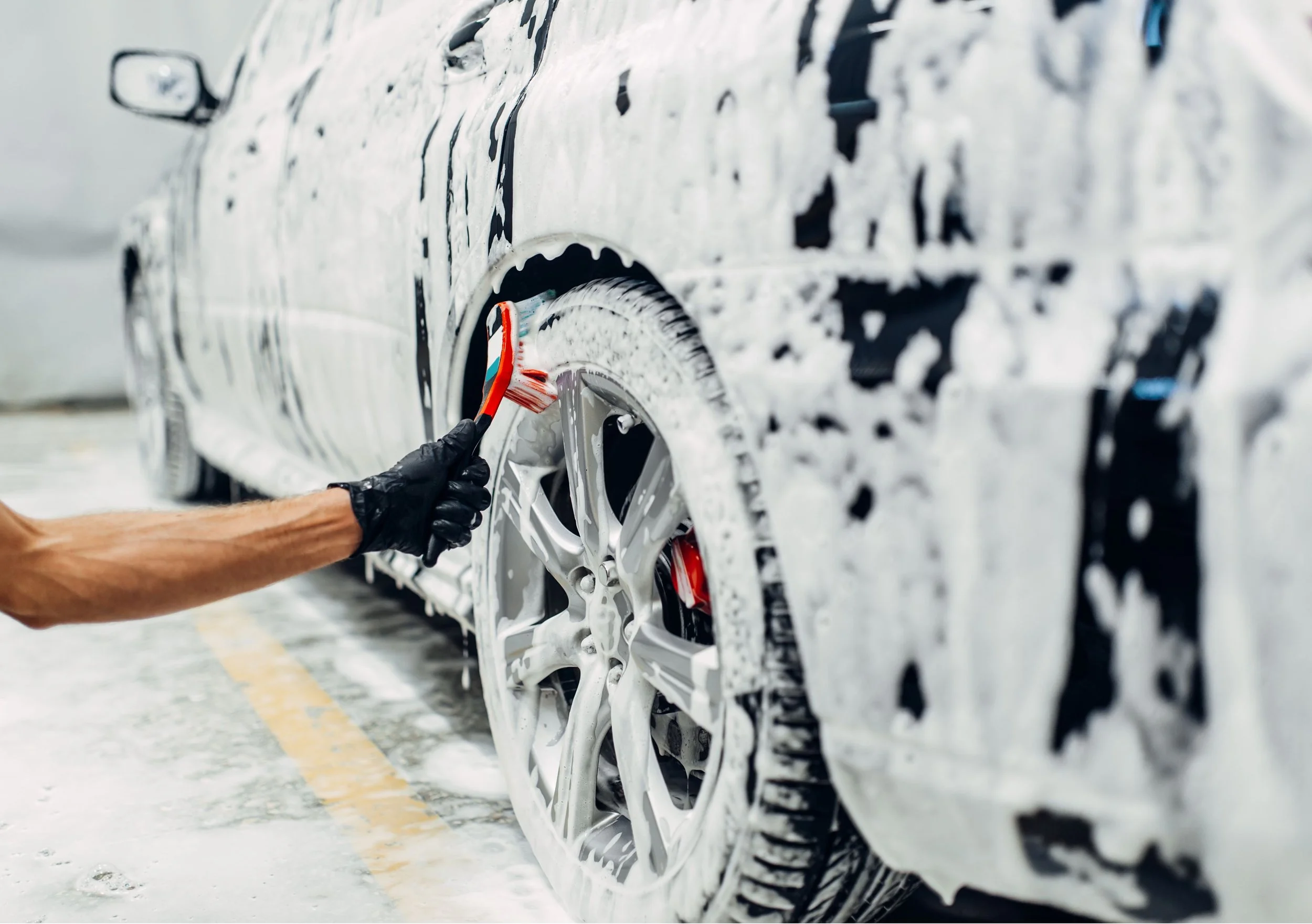 Person washing a white car with a brush, covered in soap suds, in a garage or washing station.