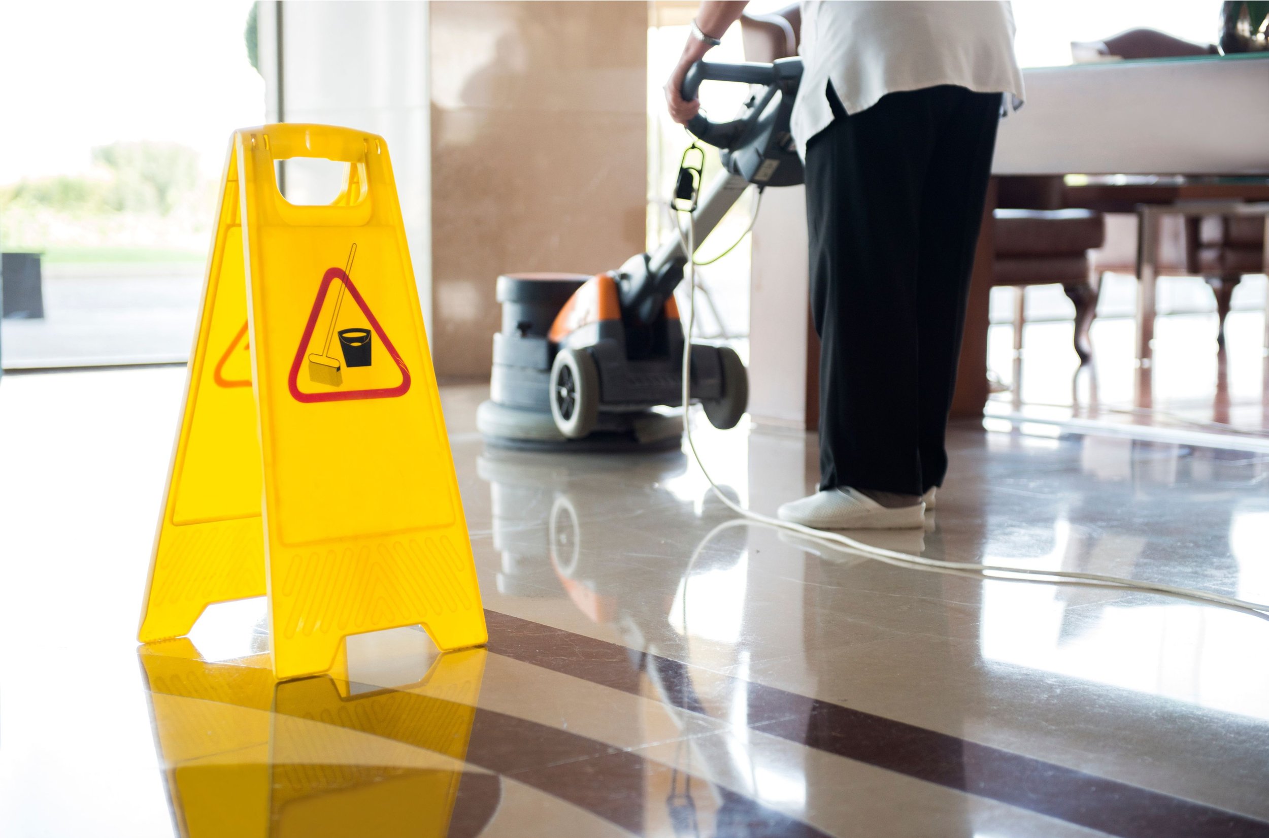 Yellow caution sign with a wet floor warning and a mop icon in front of a person cleaning the floor with a machine, near a large window and a dining area.