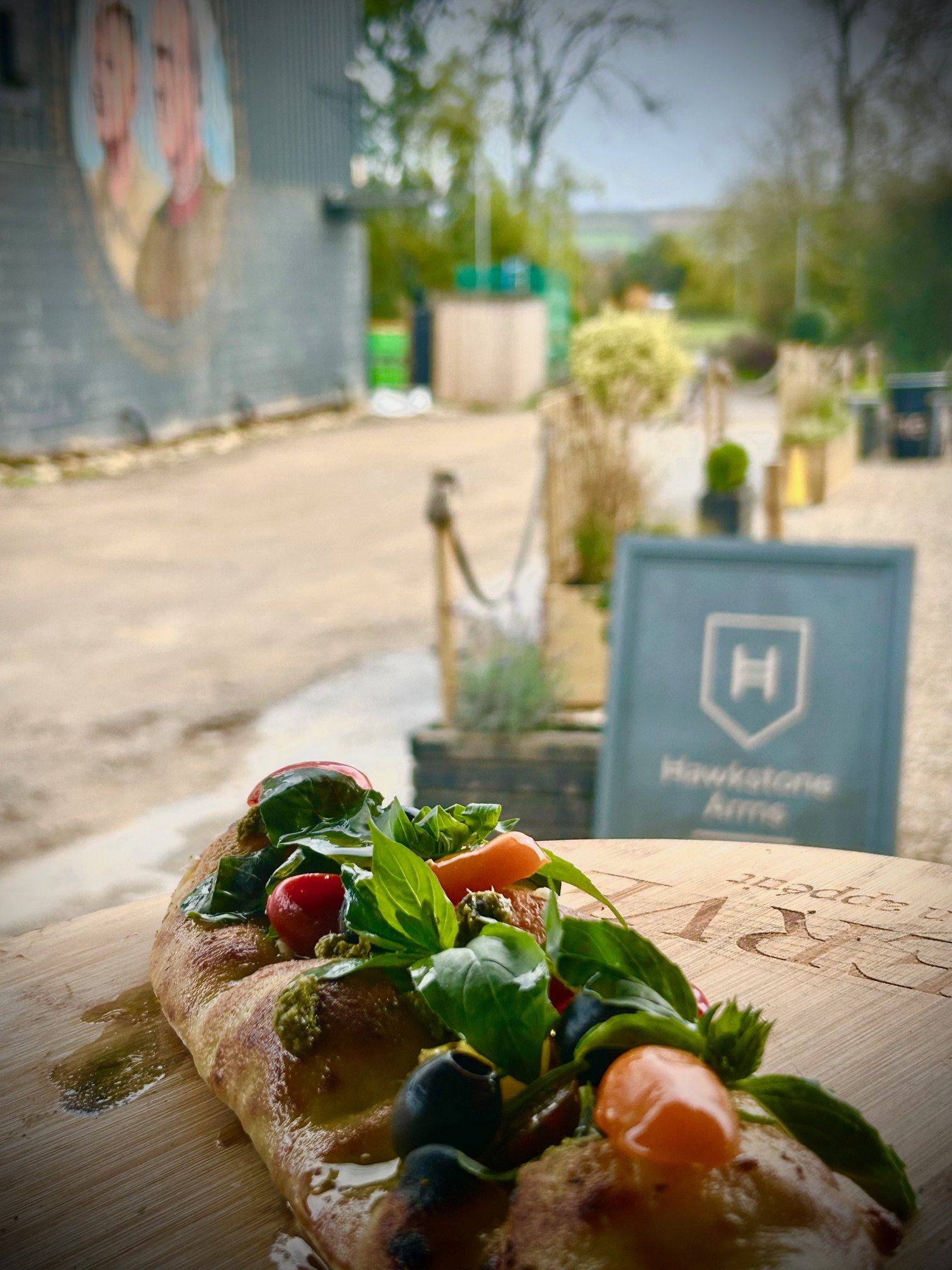 A pizza with colorful toppings including basil, cherry tomatoes, black olives, and cheese on a wooden board inside a restaurant.