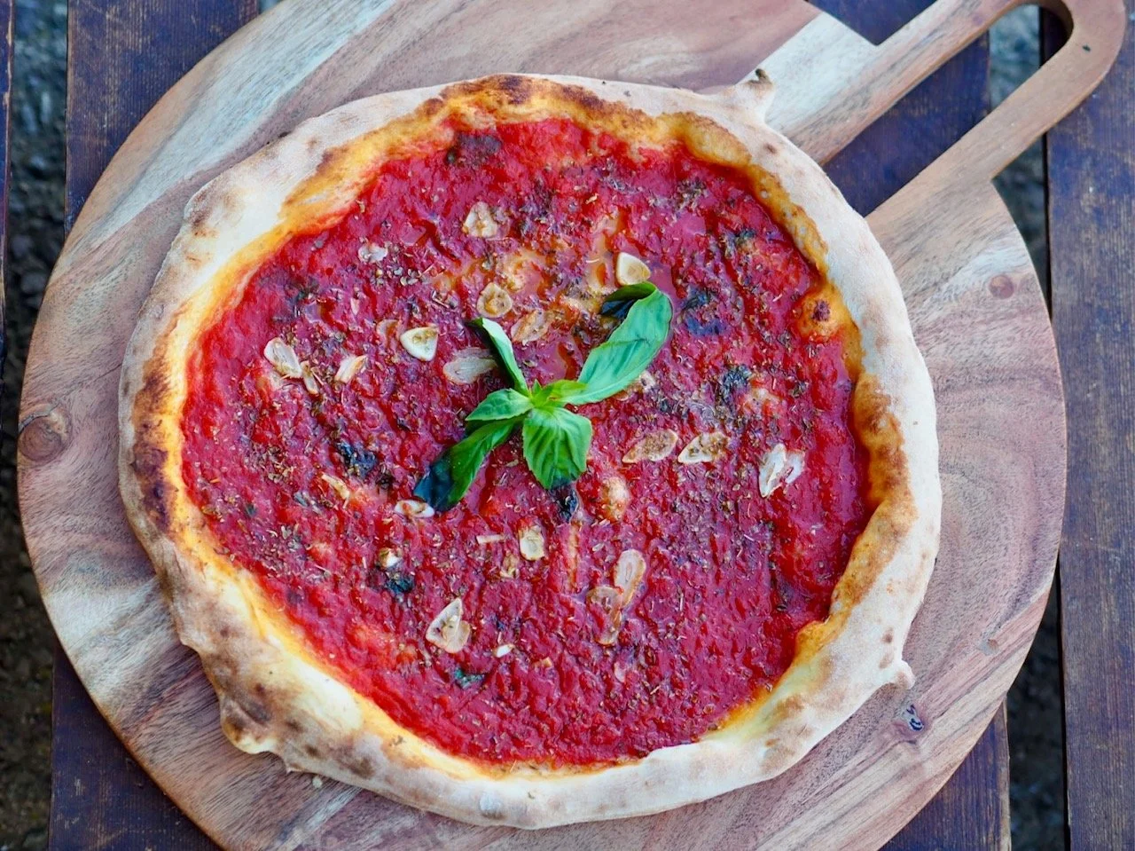 A pizza with tomato sauce, sliced garlic, and basil leaves on top, served on a wooden pizza peel.