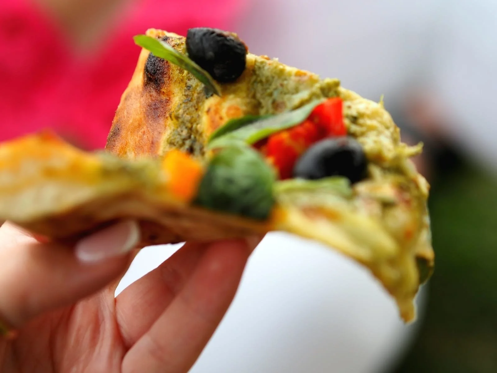 Close-up of a hand holding a slice of pizza with toppings including black olives, green peppers, and red peppers.