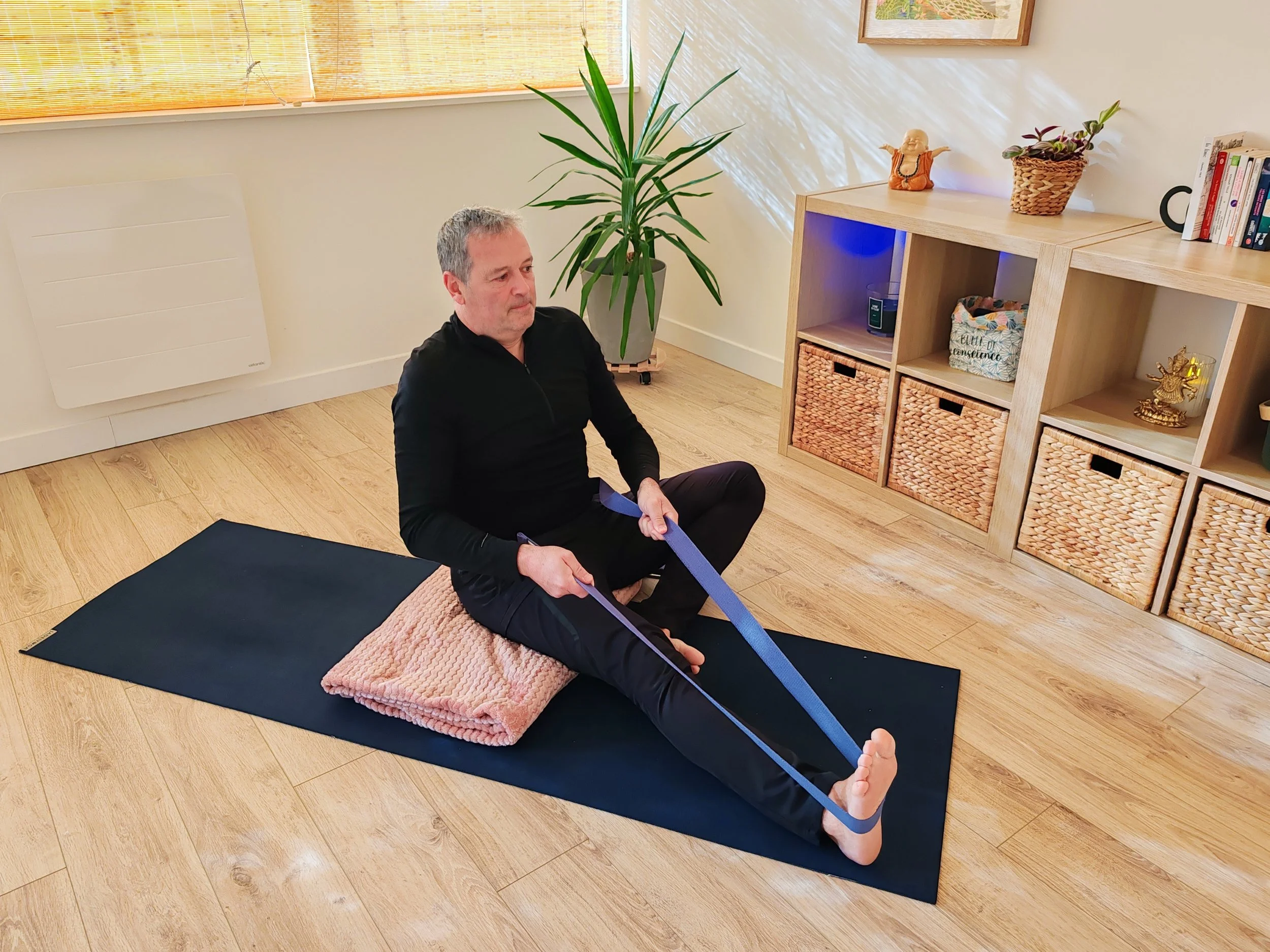Homme pratiquant une séance de stretching sur un tapis dans une pièce bien éclairée, avec des étagères en bois et des décorations naturelles autour.