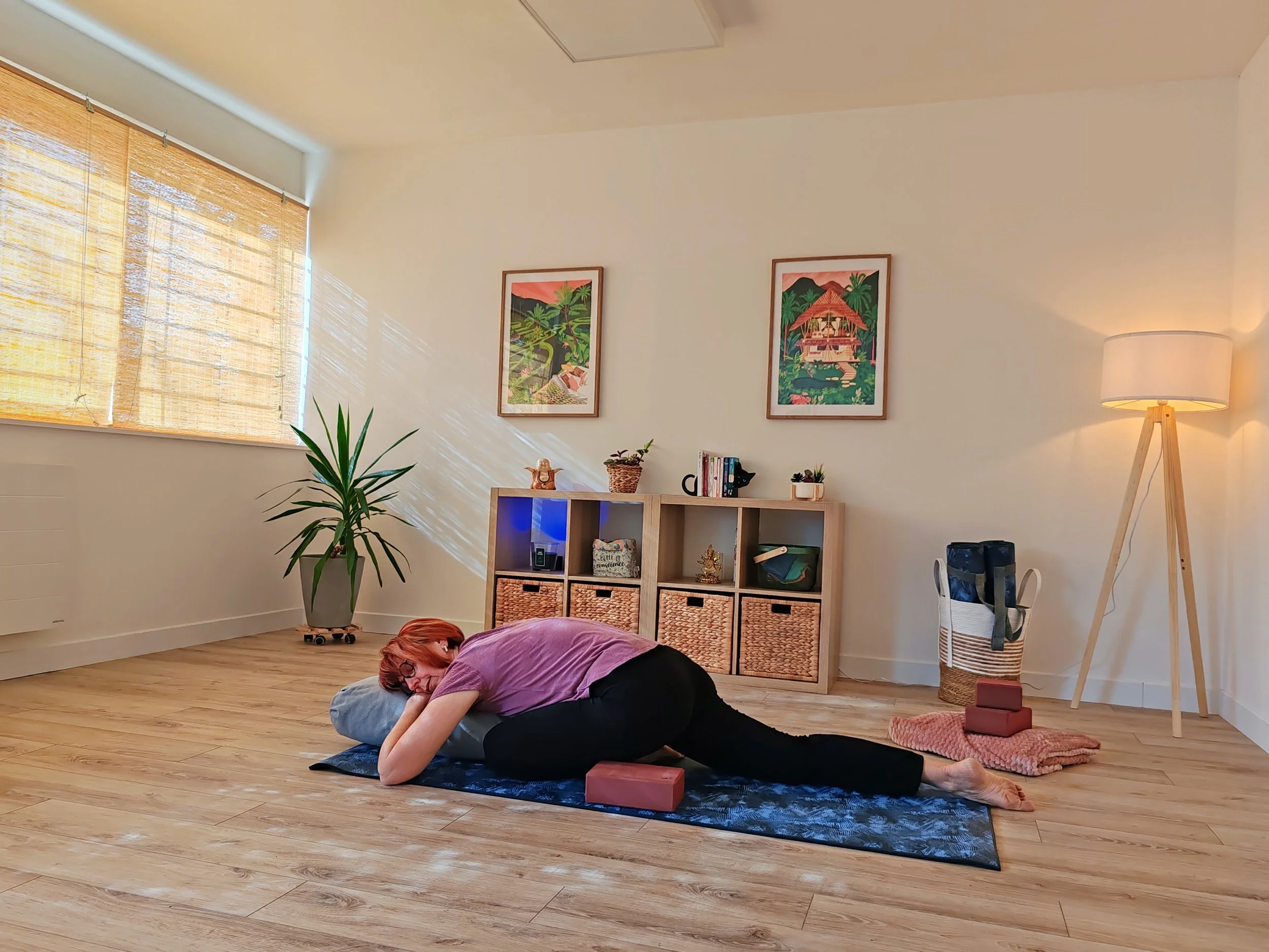 Une personne pratique le yoga, allongée sur un tapis dans une pièce bien éclairée avec une grande fenêtre, des plantes, et une étagère avec des décorations.