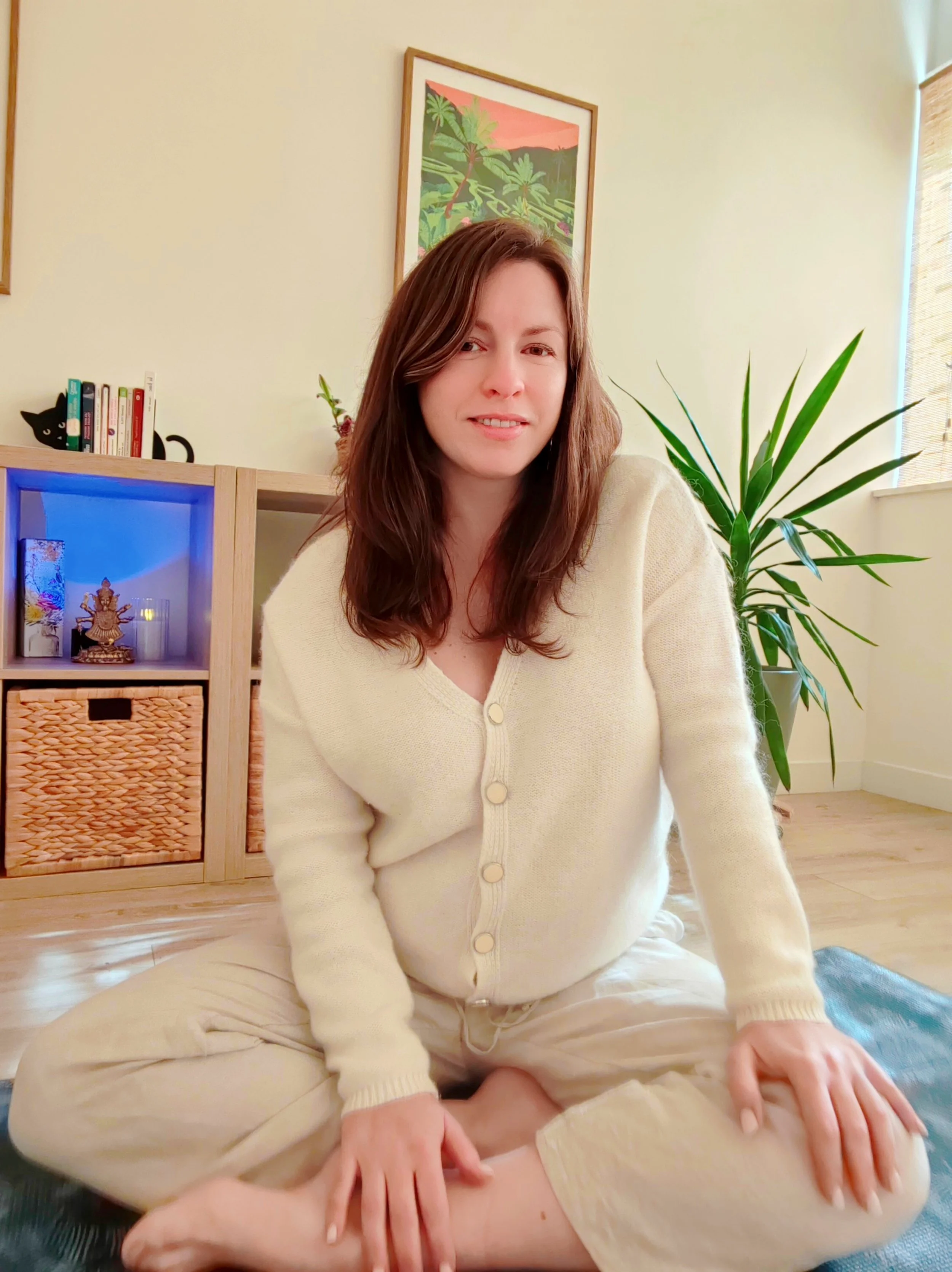Femme assise en tailleur dans une pièce avec des plantes et des étagères.