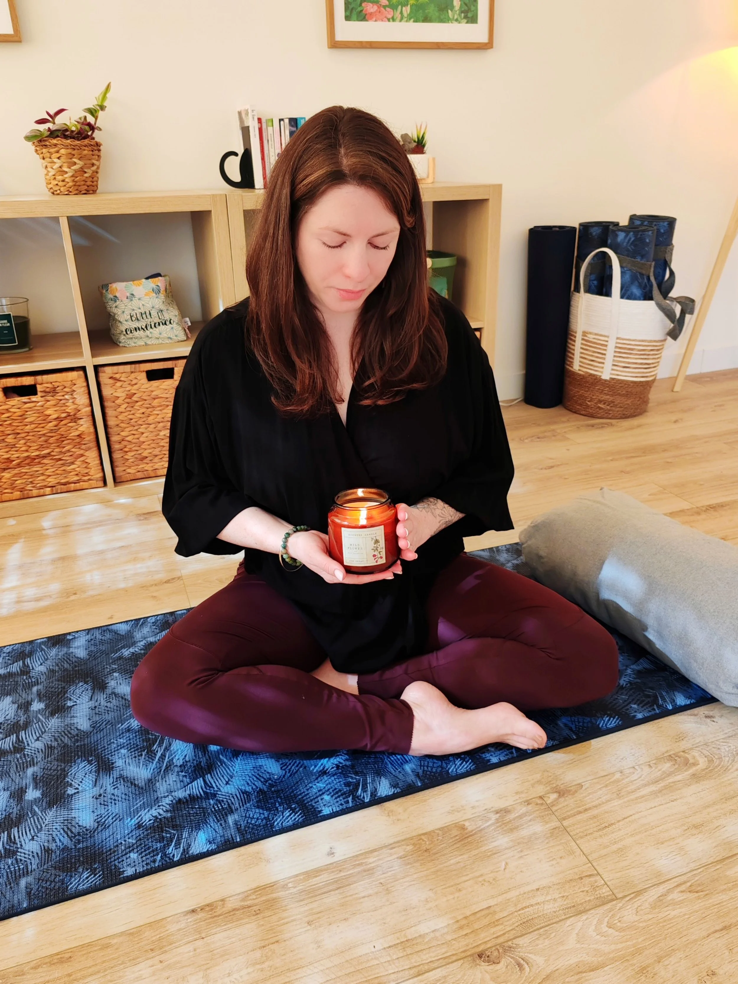 Une femme assise en méditation sur un tapis, tenant une bougie allumée dans ses mains, dans une pièce chaleureuse et bien décorée.