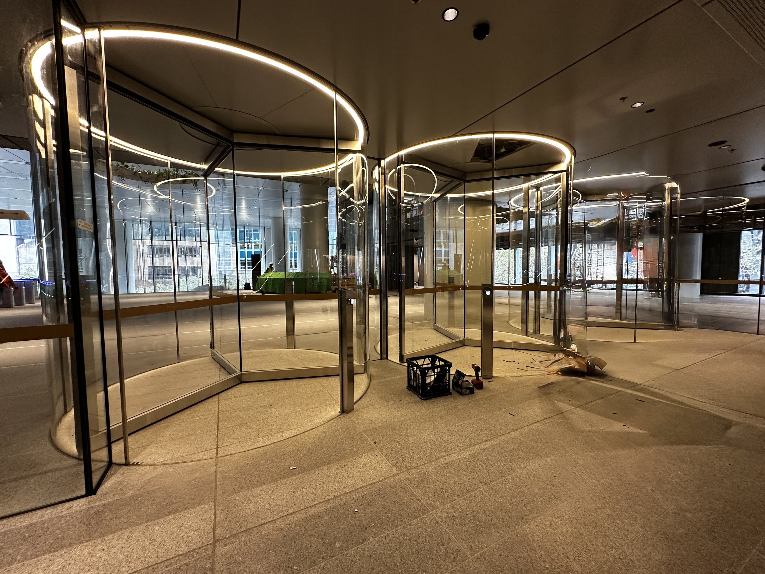 Interior of a modern building with glass revolving doors, illuminated ceiling lights, large windows, and construction tools on the floor.