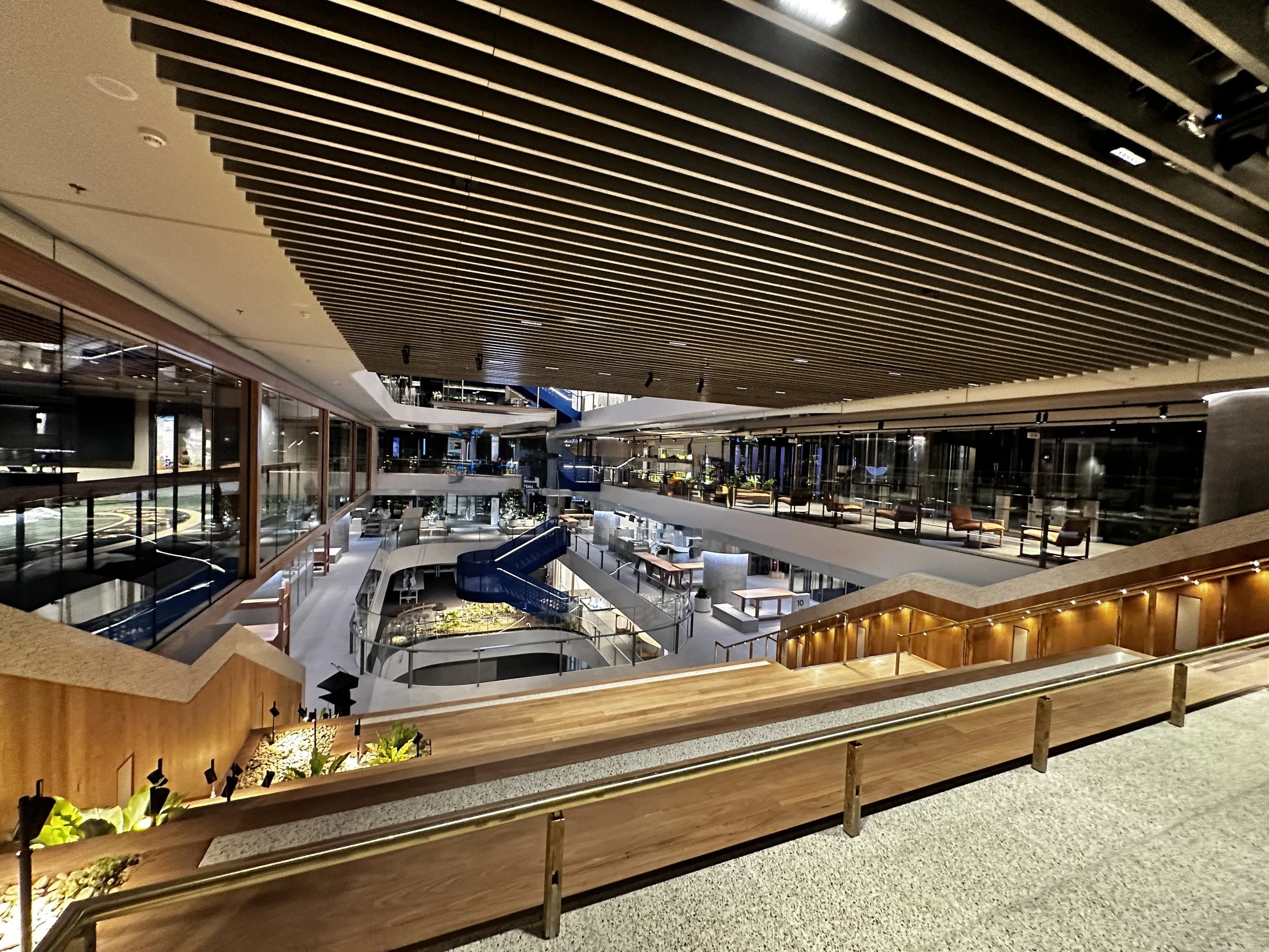 Interior view of a modern multi-level shopping mall with wooden accents, glass storefronts, and escalators connecting floors.