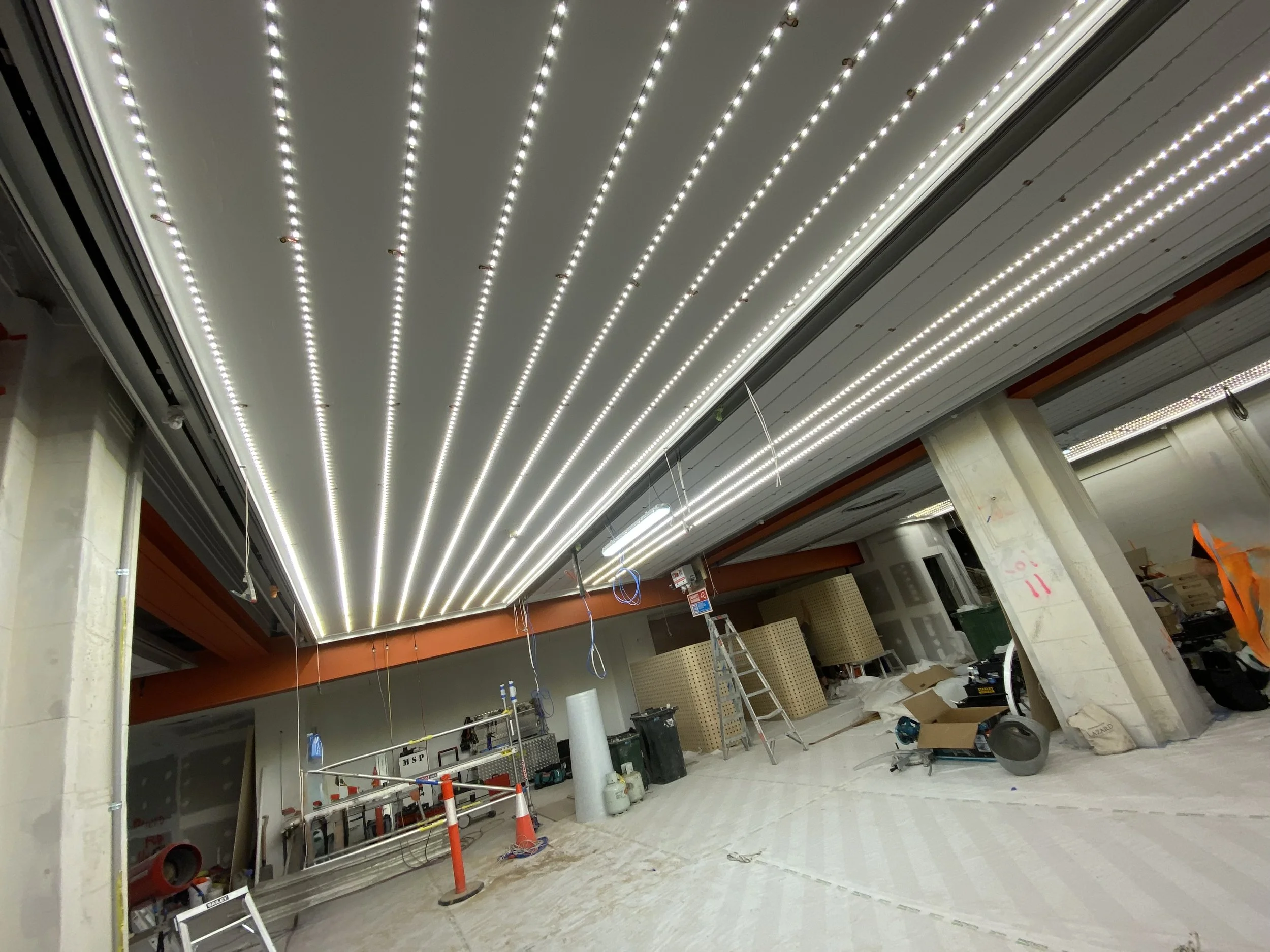 Indoor construction site with exposed ceiling featuring rows of LED strip lights, scaffolding, tools, and various construction materials scattered around.