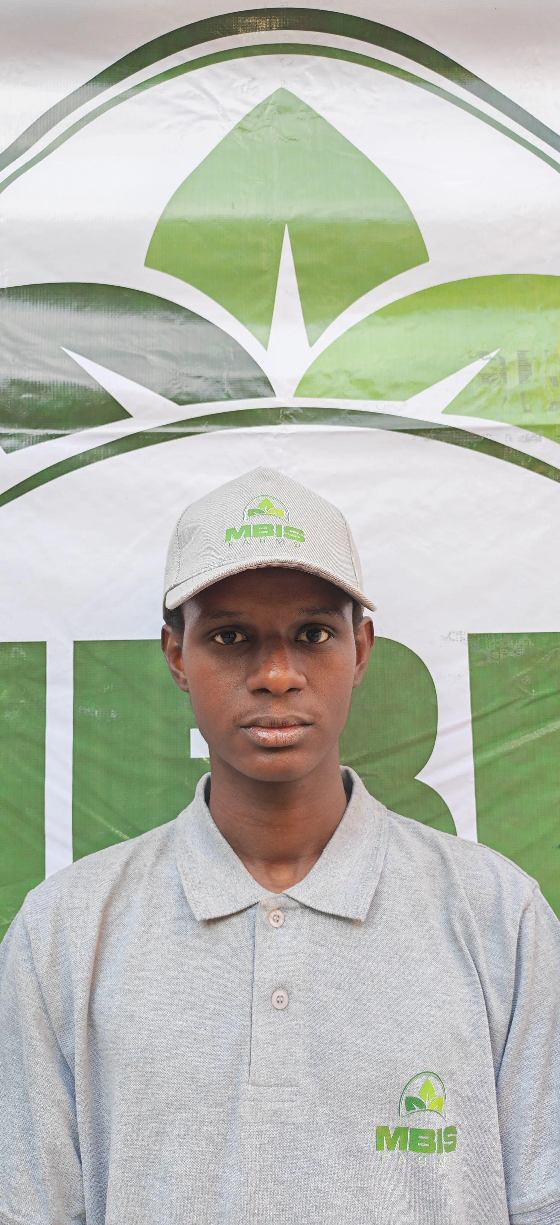 Young man wearing gray polo shirt and baseball cap with 'MBIS Farm' logo, standing in front of a green and white MBIS Farms banner.