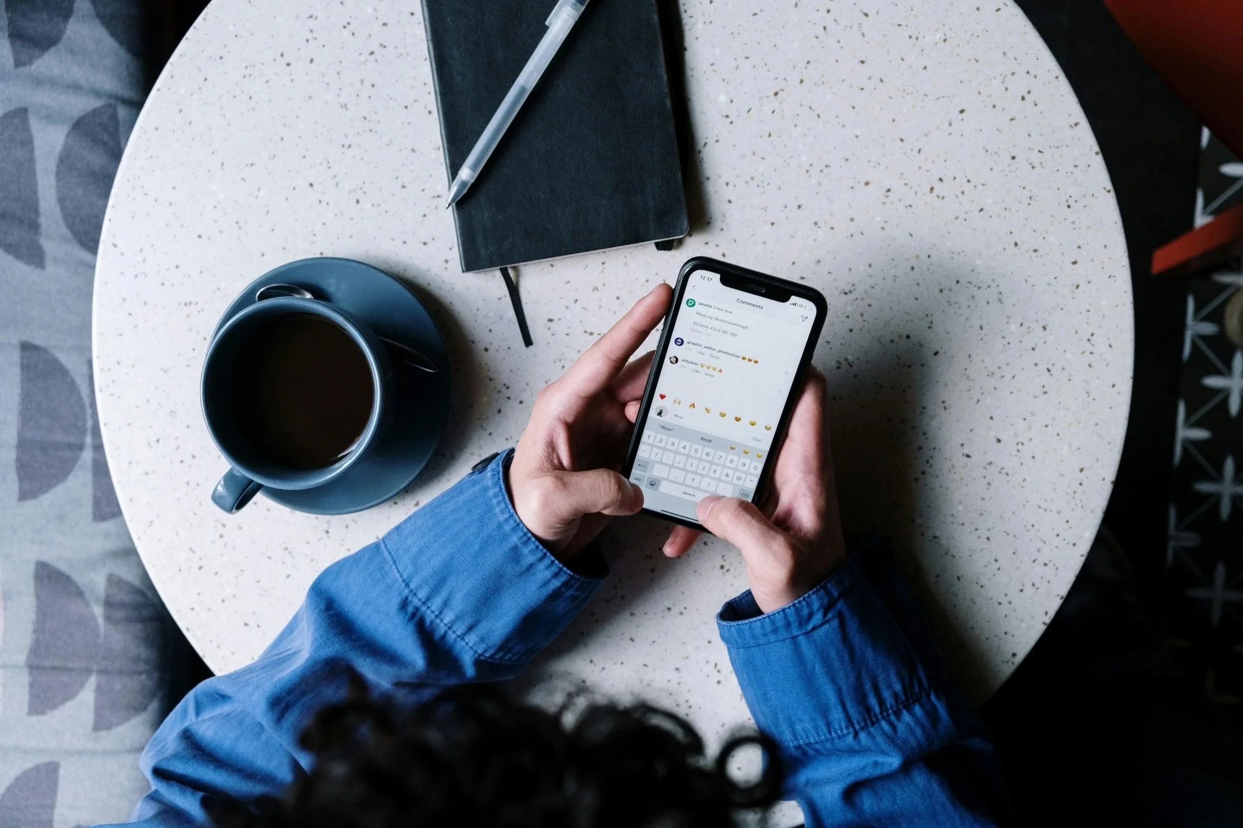 Person in a blue jacket sitting at a round, speckled table, looking at their phone. On the table, there is a black notebook with a pen, a blue mug filled with coffee, and a smaller blue saucer.