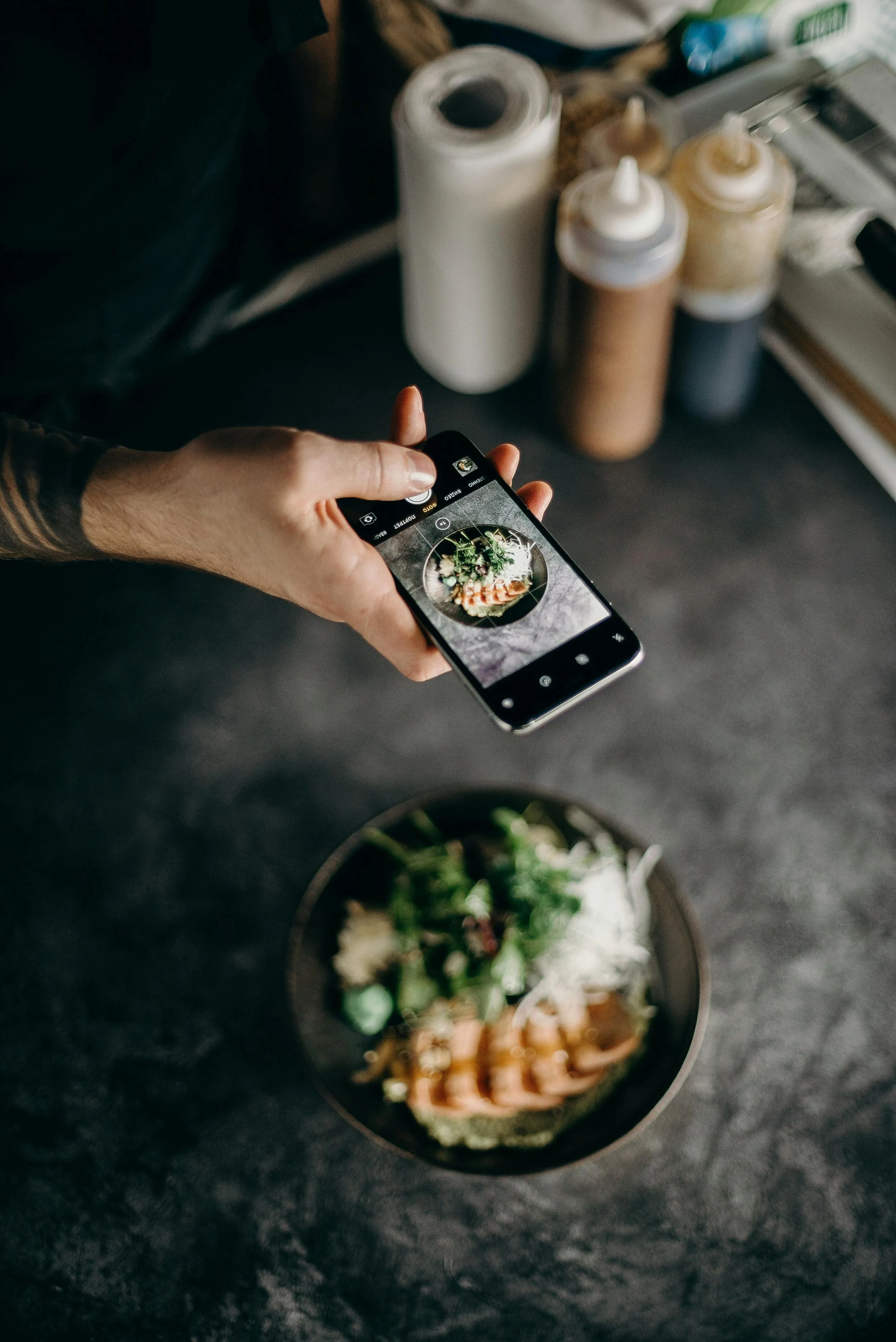 Person taking a photo of a bowl of food with a smartphone in a kitchen or cafe setting, with condiment bottles in the background.