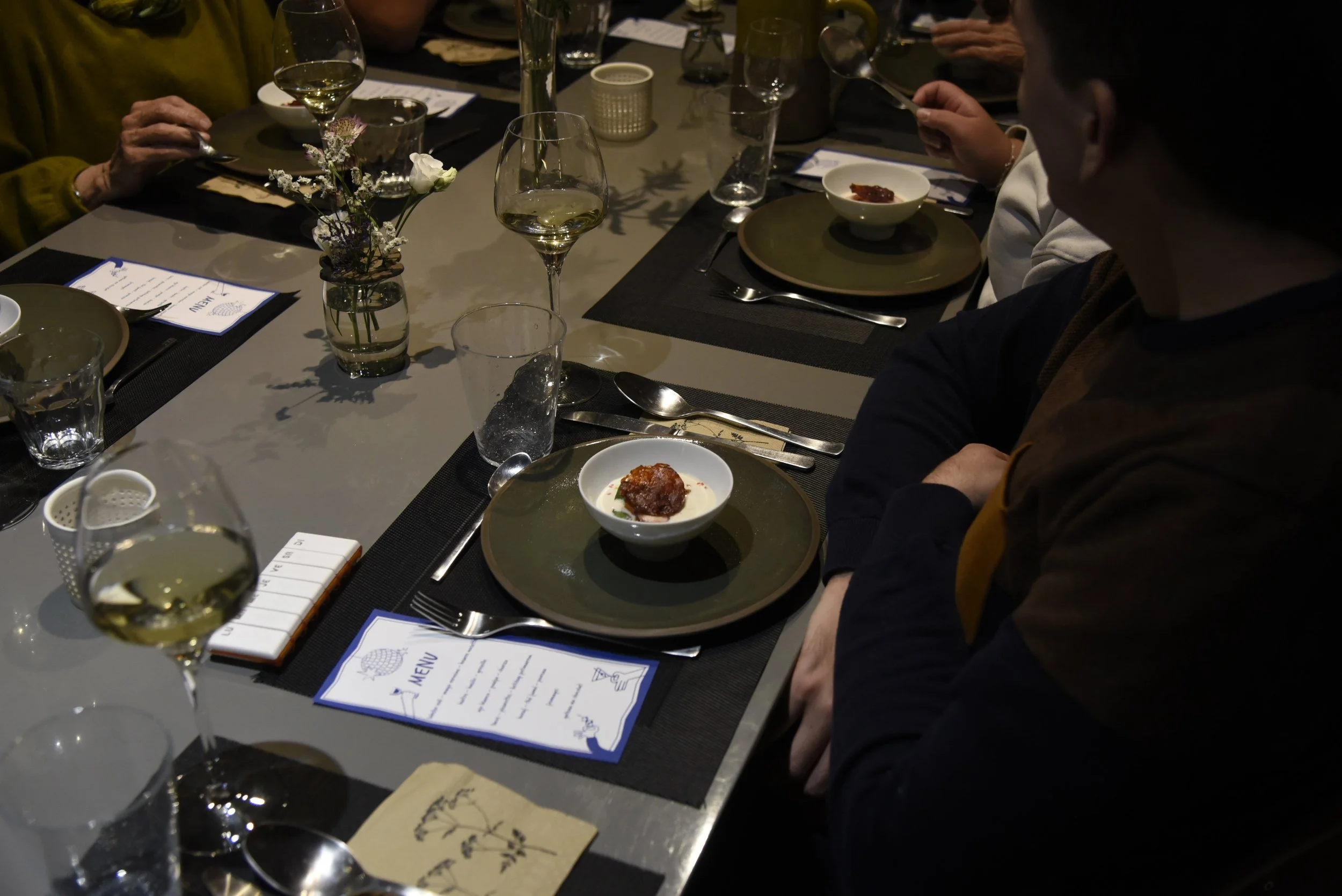 Une table mise pour un repas avec des assiettes, des verres de vin blanc, des couverts, une petite décoration florale centrale, et un menu. Il y a plusieurs personnes assises autour, certaines tenant des cuillères. La table a un look élégant et moderne.