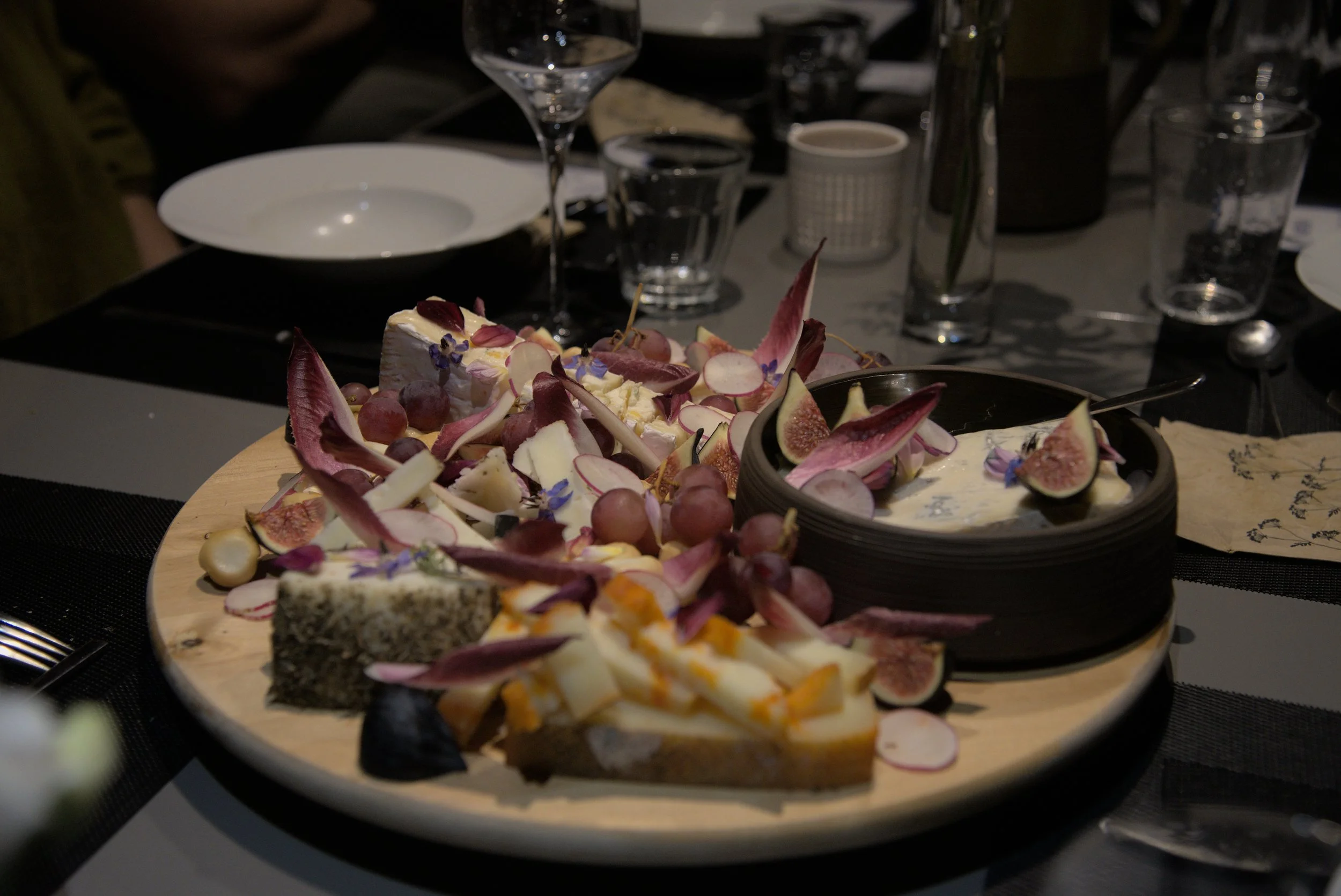 Plate de fromages et fruits avec raisins, figues, radis, fleurs comestibles, sur fond de table de dîner avec verre de vin et vaisselle.