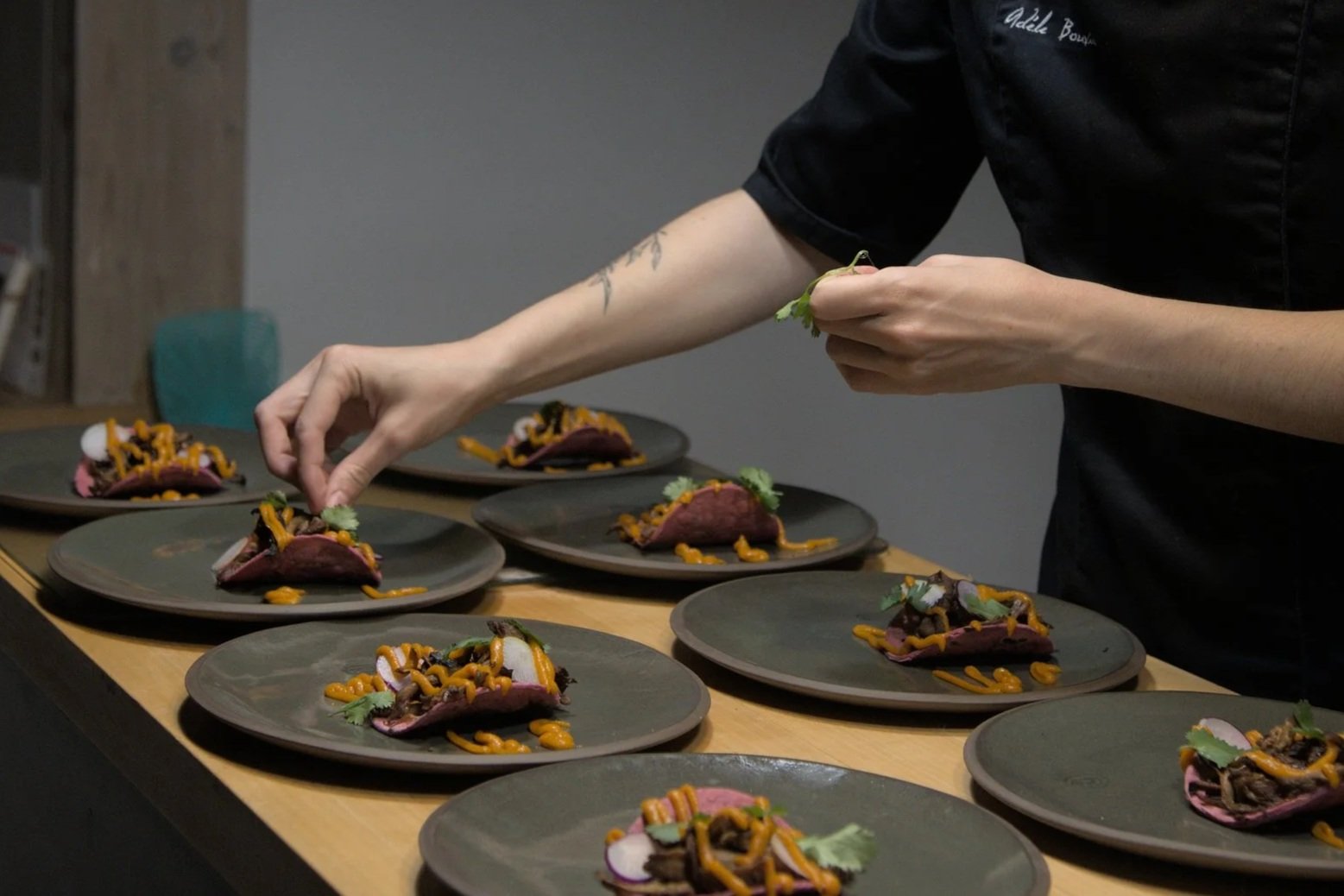 Un chef prépare une assiette de nourriture sophistiquée avec de la viande, des légumes et des sauces sur des assiettes noires, en utilisant des pincettes.