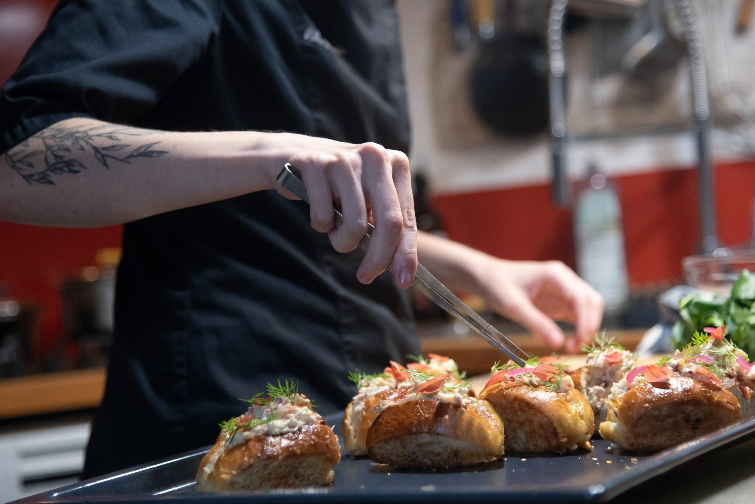 Chef préparant des petits pains garnis à l'aide d'une pince, dans une cuisine professionnelle.