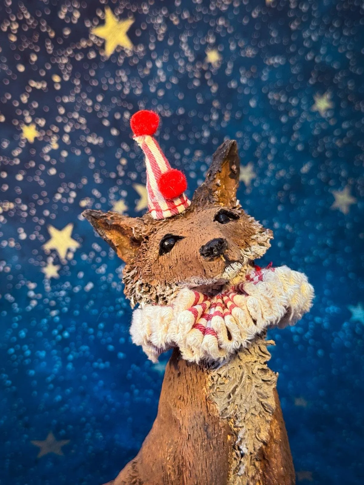 A decorative figure of a fox made of clay, adorned with a ruffled collar and wearing a small party hat with pom-poms, against a background of blue with star shapes and bokeh lights.