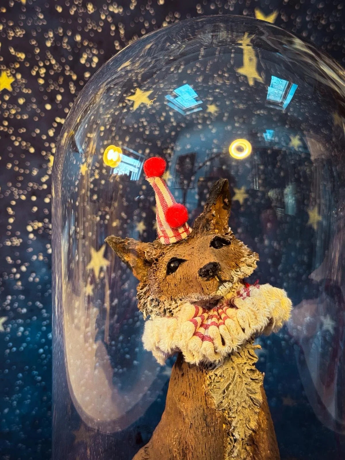Decorative fox figurine with a circus hat and scarf inside a glass sphere, with star-shaped and circular lights in the background.