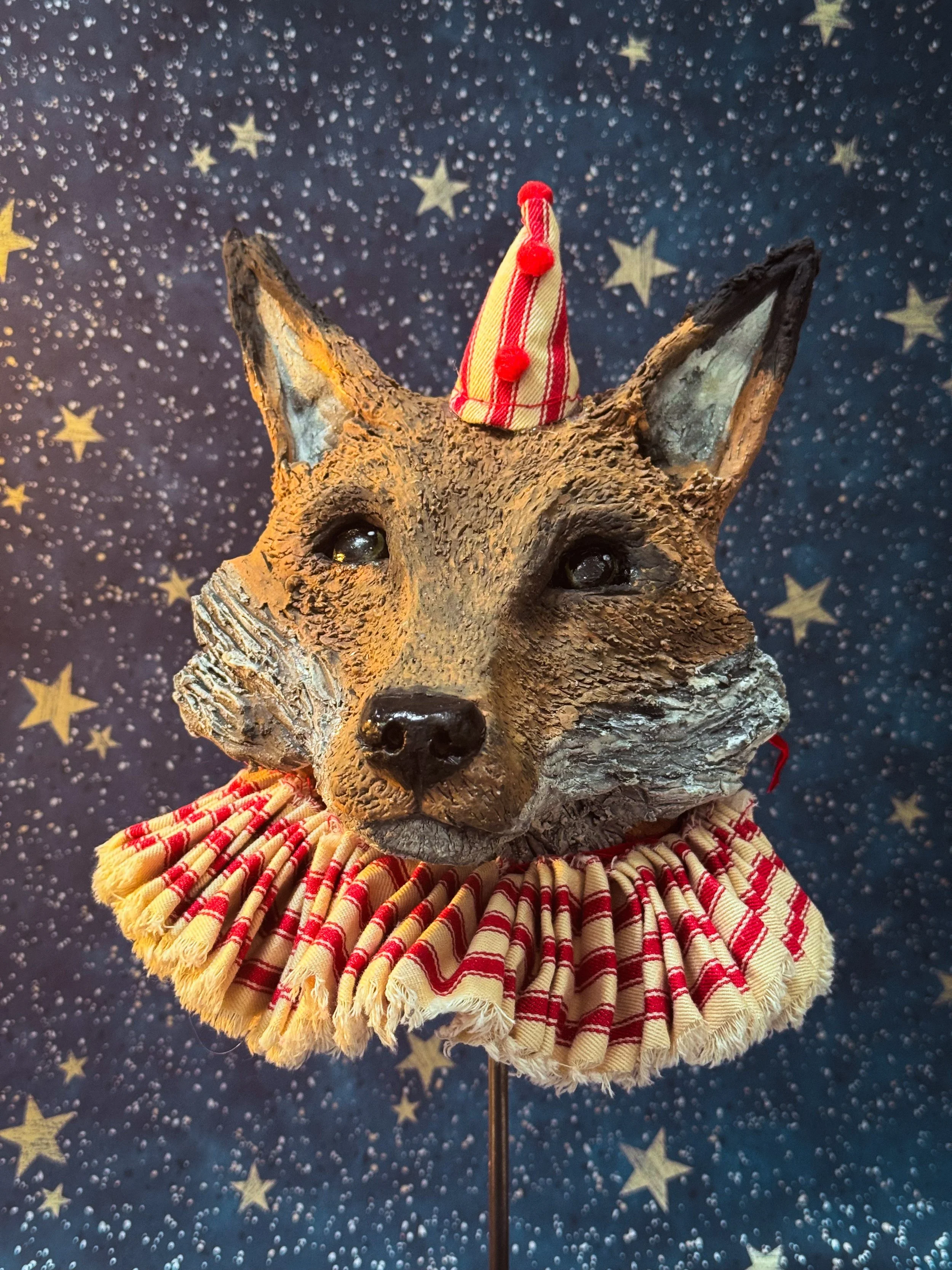A fox with a clown hat on top, dressed in yellow and red striped fabric, and an orange and red ruffled collar, against a background with yellow stars and white dots.