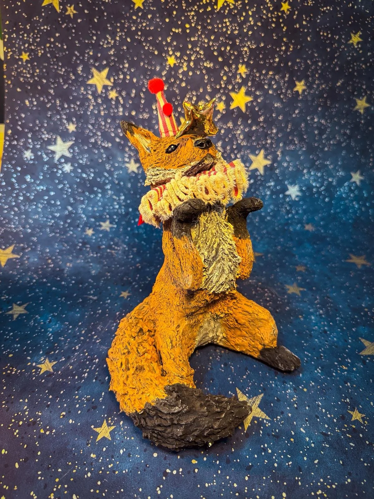 A decorative sculpture of a fox standing on its back legs with its front paws together, wearing a small crown, a striped scarf, and a star-shaped balloon. The background features a starry pattern with gold and white stars on a dark blue backdrop.